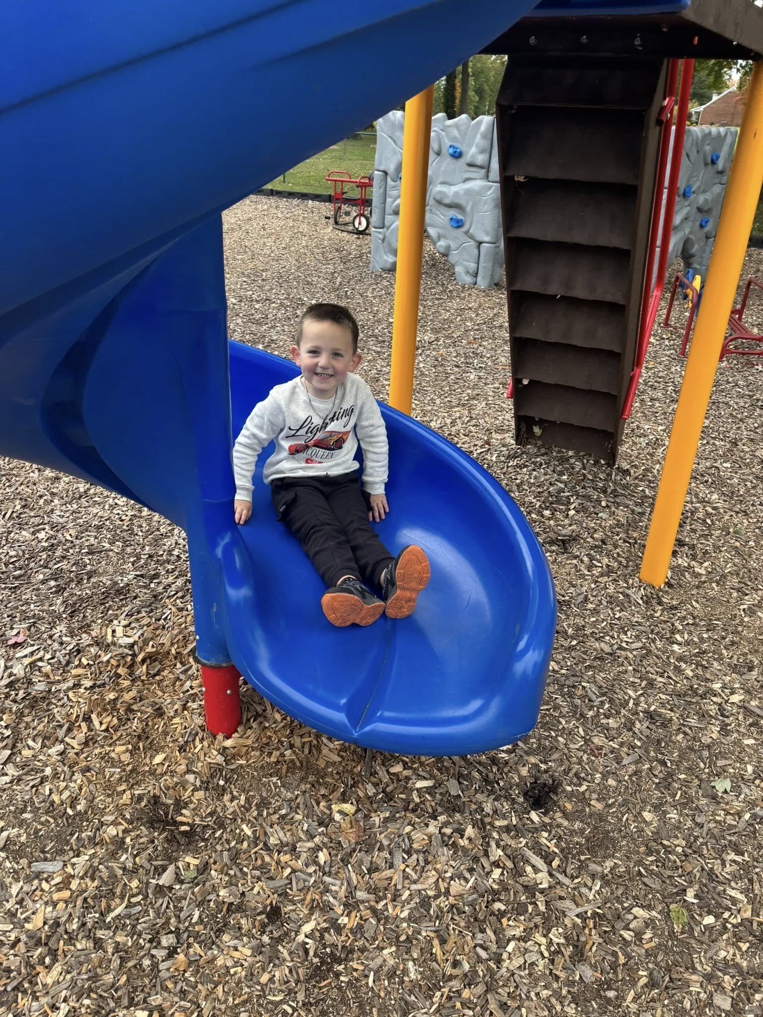 A young boy sitting at the bottom of a blue playground slide, smiling, at a playground covered with wood chips, with a climbing wall and other playground equipment in the background.