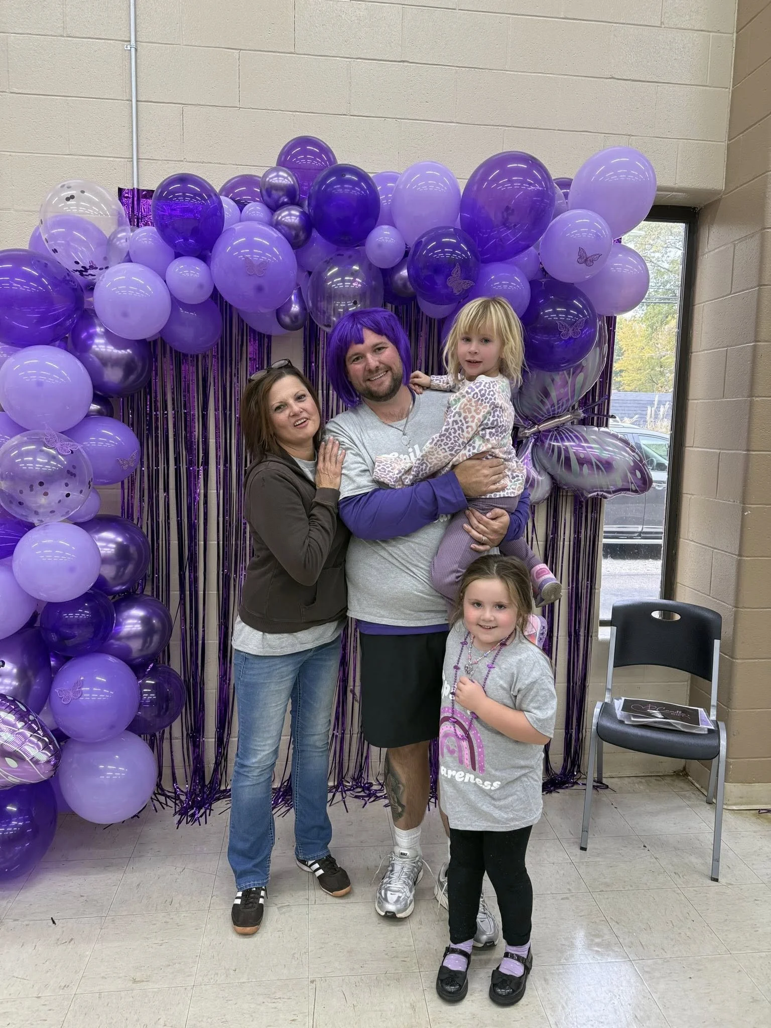 A group of four people, including two young girls, posing together in front of purple balloon decorations, with a purple backdrop and butterfly accents, inside a room near a window.