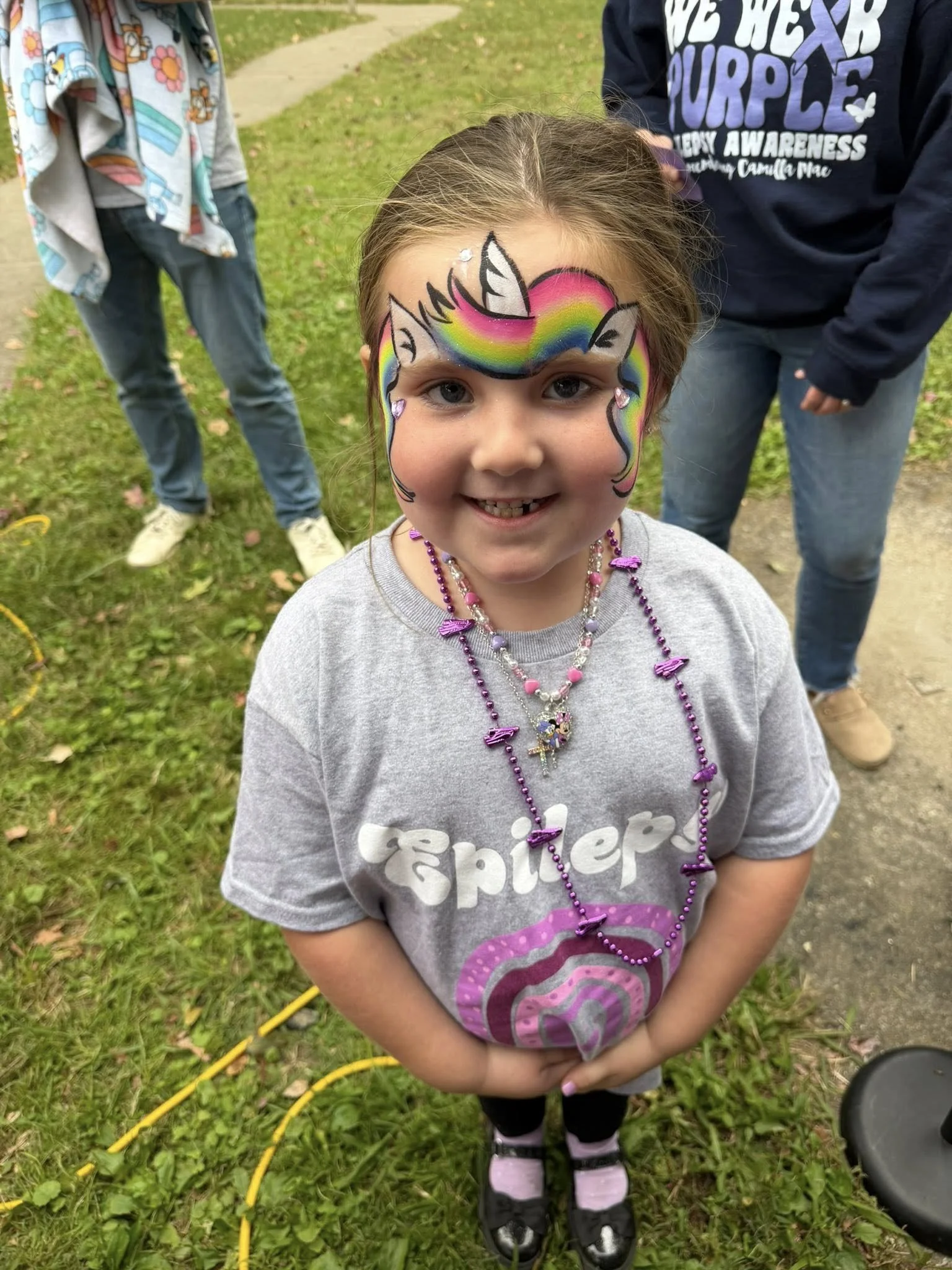Young girl with colorful unicorn face paint, smiling, wearing a gray T-shirt with a lollipop graphic, purple beaded necklaces, standing outdoors on grass with two other children nearby.