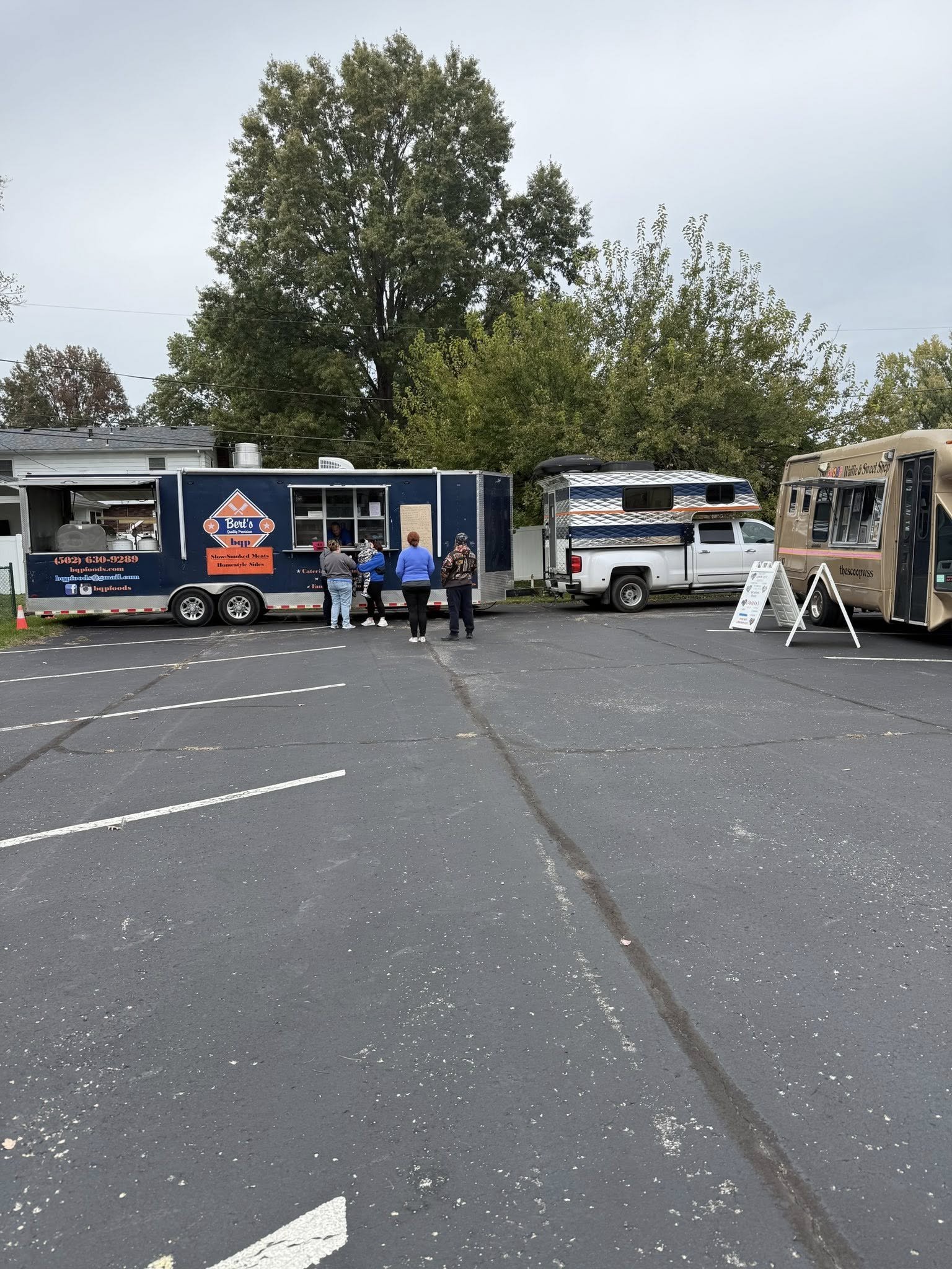 Food trucks parked in a parking lot with people lining up at one of the trucks, trees in the background, and a cloudy sky.