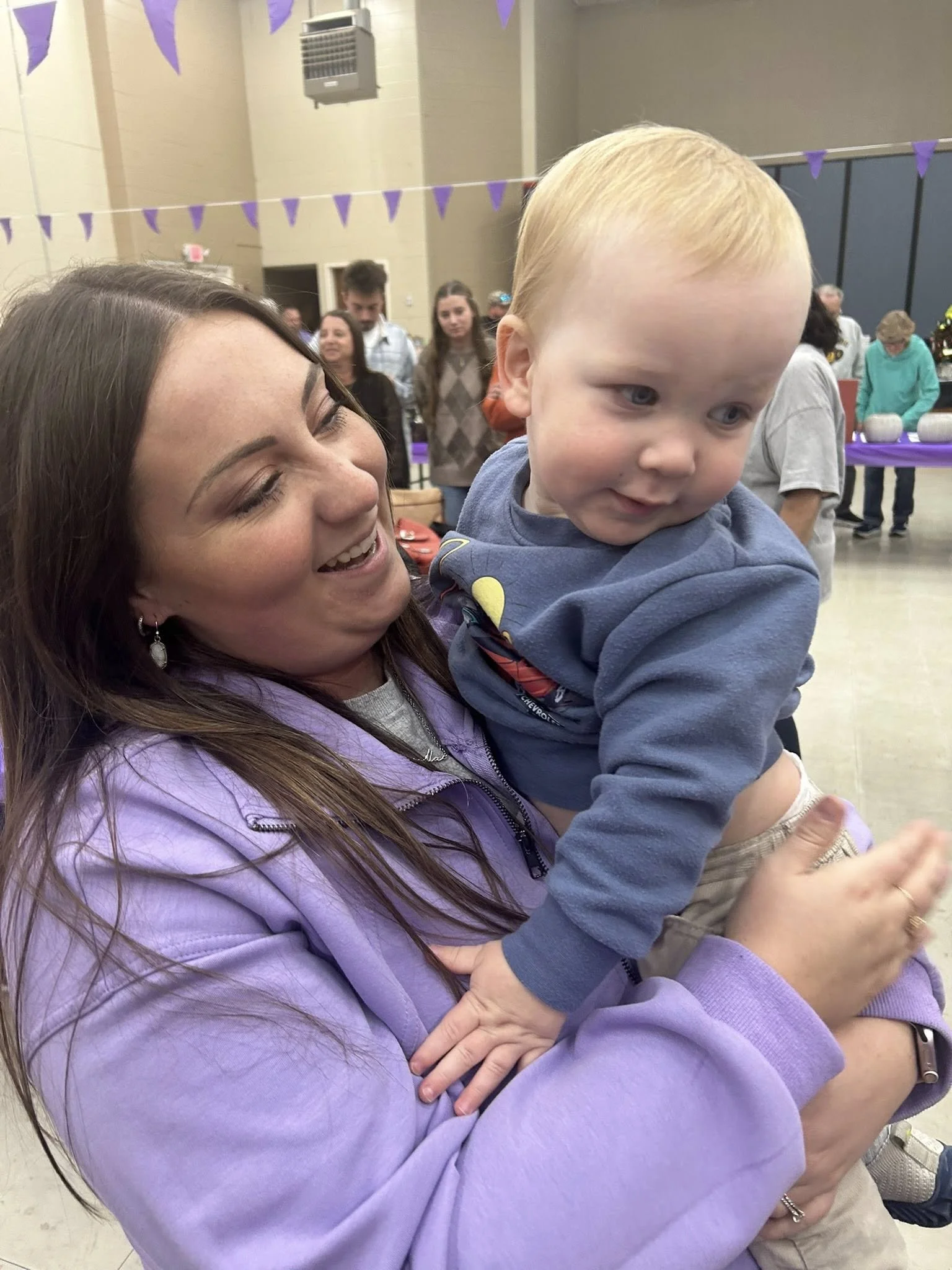 A woman with long brown hair holding a young blond boy, both smiling, at an indoor event decorated with purple banners and people in the background.
