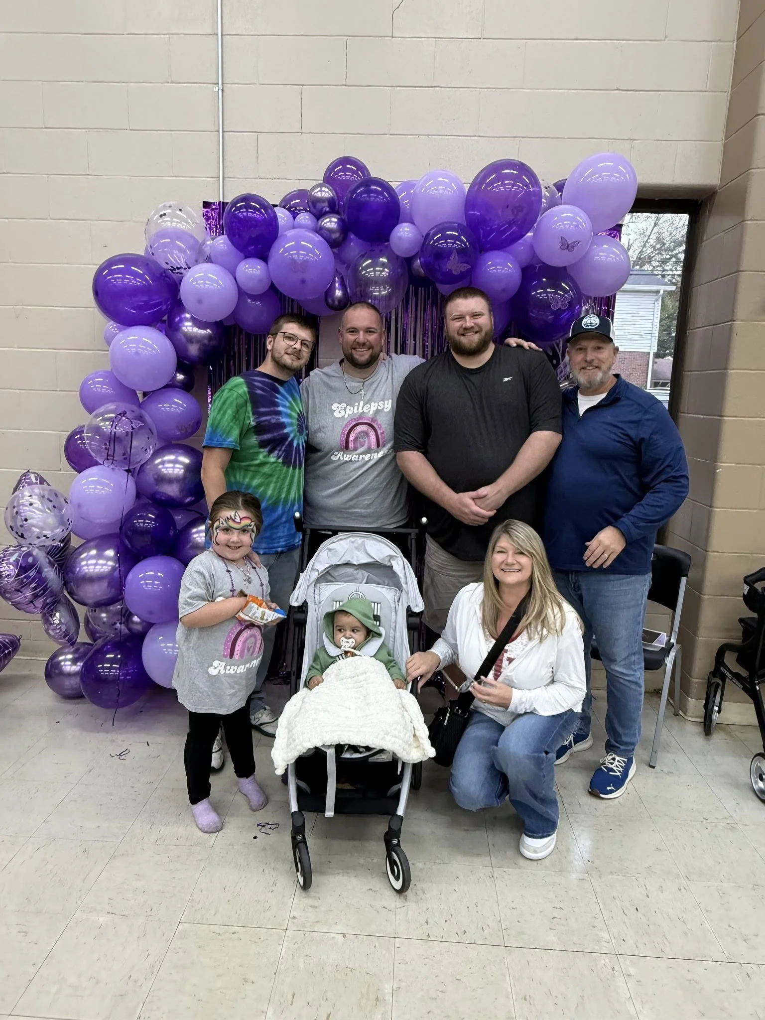 A group of six people and two children posing in front of purple balloons with butterfly decorations. One child is in a stroller, and the other has face paint and a snack. They are indoors, in a celebration setting.