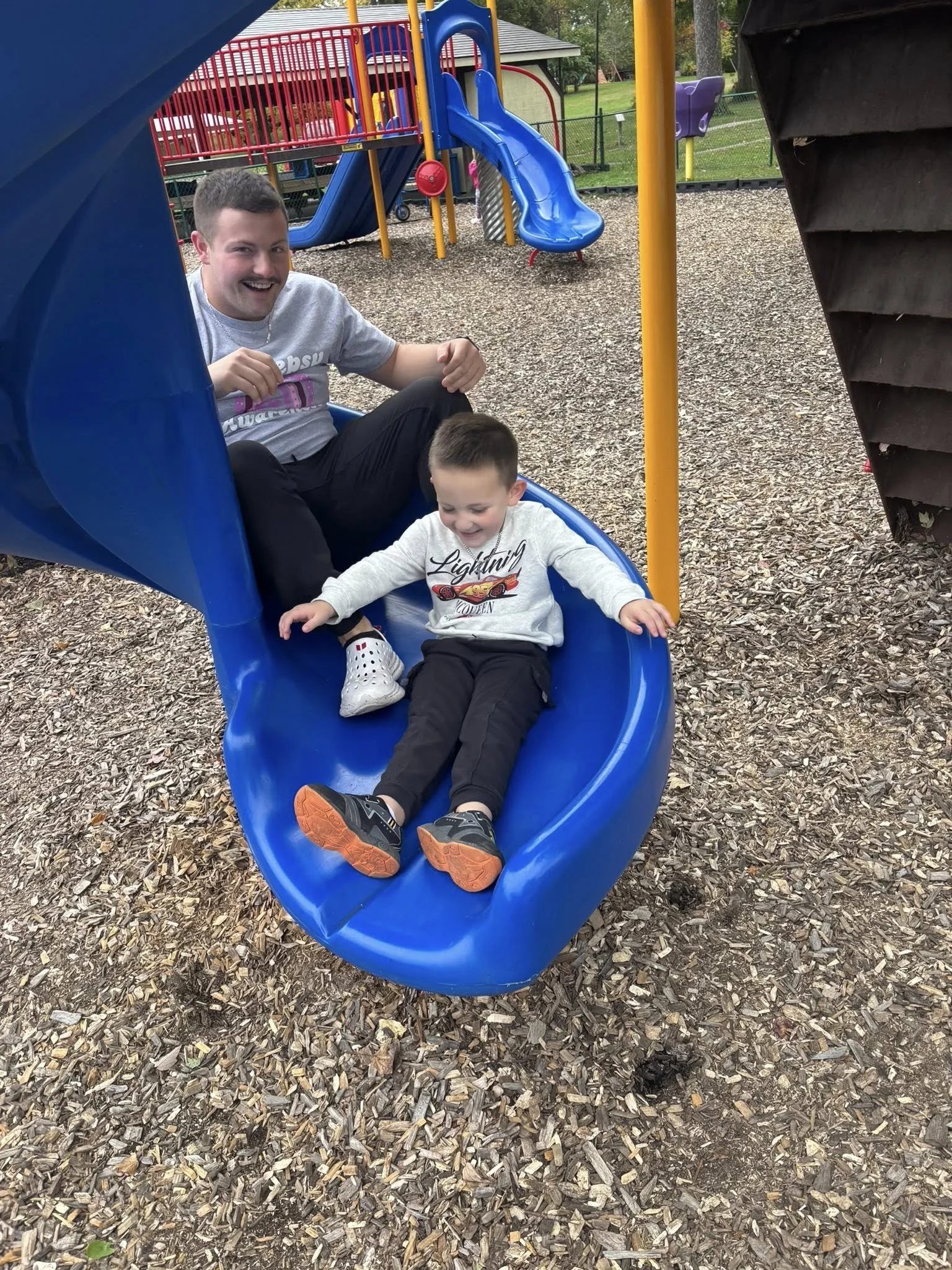 Two children and an adult on a blue slide at a playground, surrounded by wood chips.