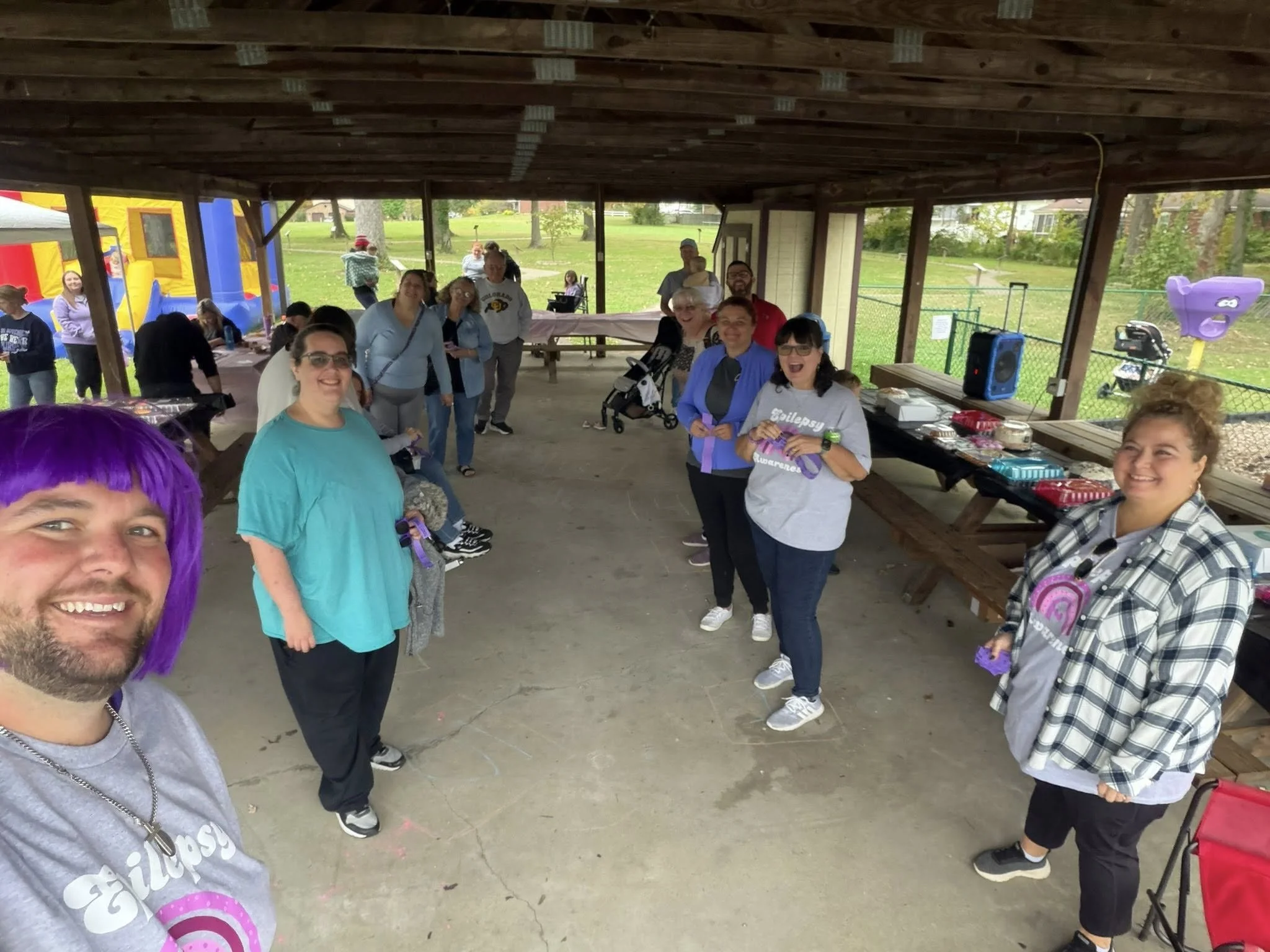 Group of people in a pavilion during an outdoor event. People are smiling, some holding purple lanyards. Behind them are tables with food and items, and a fenced sports field is visible outside.