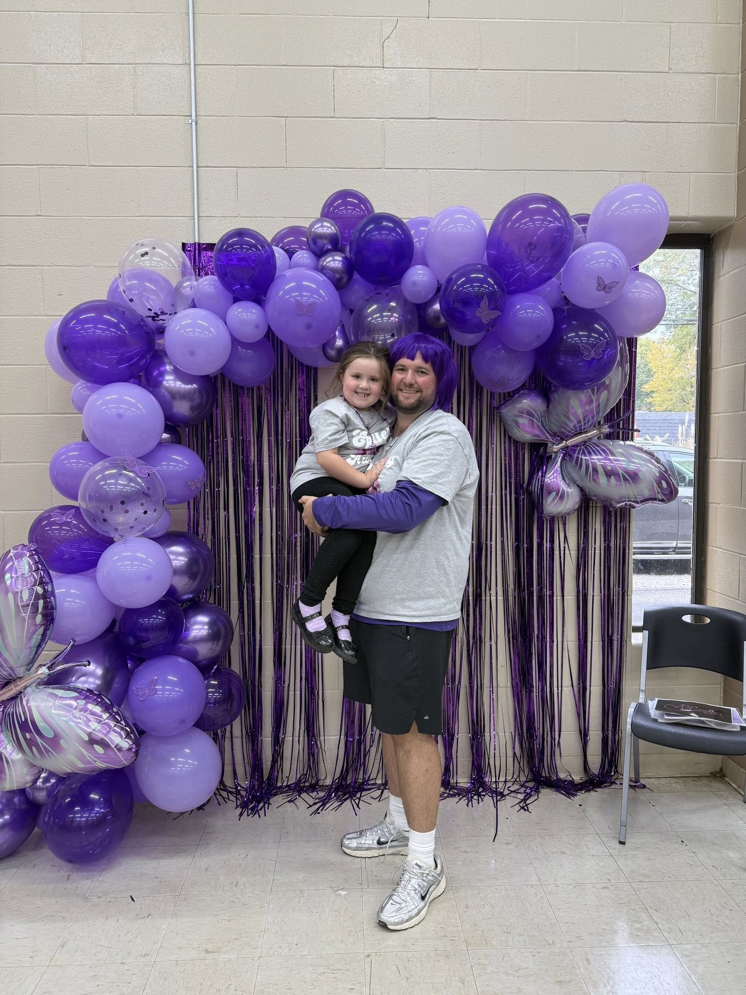 A man and a young girl smile for a photo in front of a purple balloon arch with butterfly and polka dot balloons, with purple metallic streamers hanging behind them, inside a room with a beige brick wall and a window showing outdoor trees and parked 