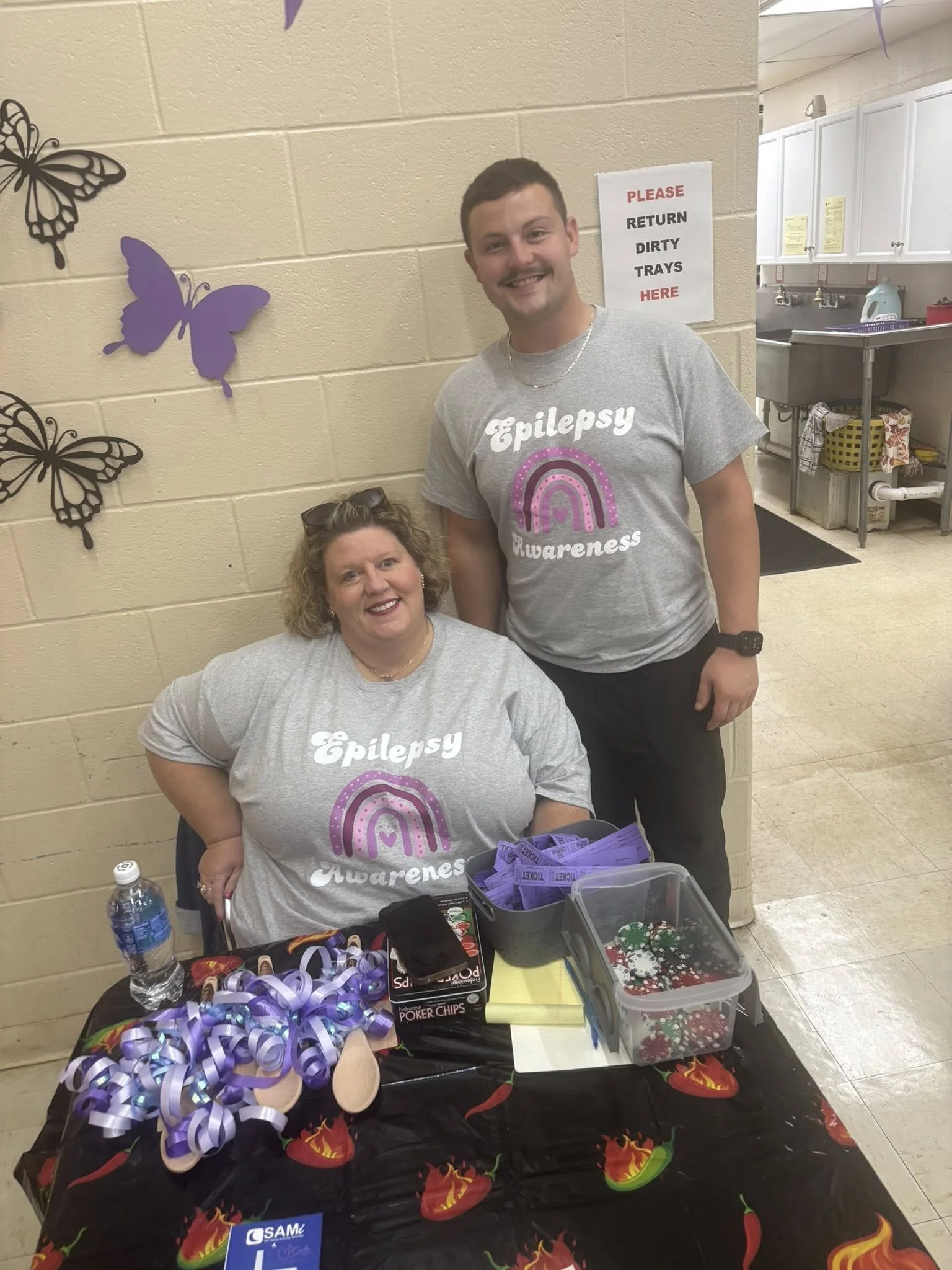 A woman and a man wearing Epilepsy Awareness t-shirts at a table with purple ribbons, poker chips, and giveaway items, standing in an indoor setting with butterfly decorations on a beige wall and a sign requesting return of dirty trays.
