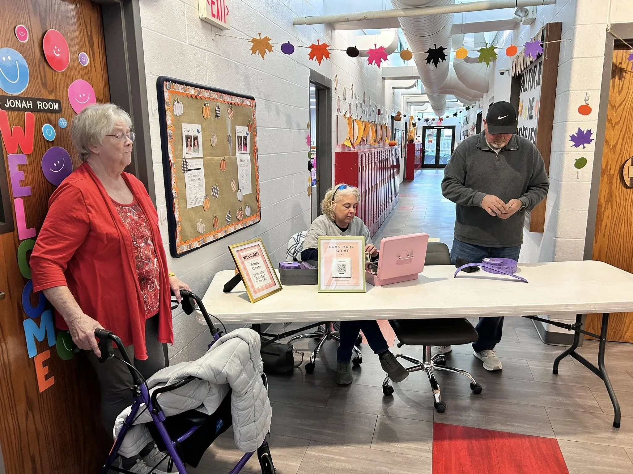 A woman in a red jacket stands next to a walker with a white quilted bag, at a check-in table. Two other women and a man are seated at the table, which has a pink payment scanner, sign-in sheets, and a welcome sign. The hallway is decorated with autu