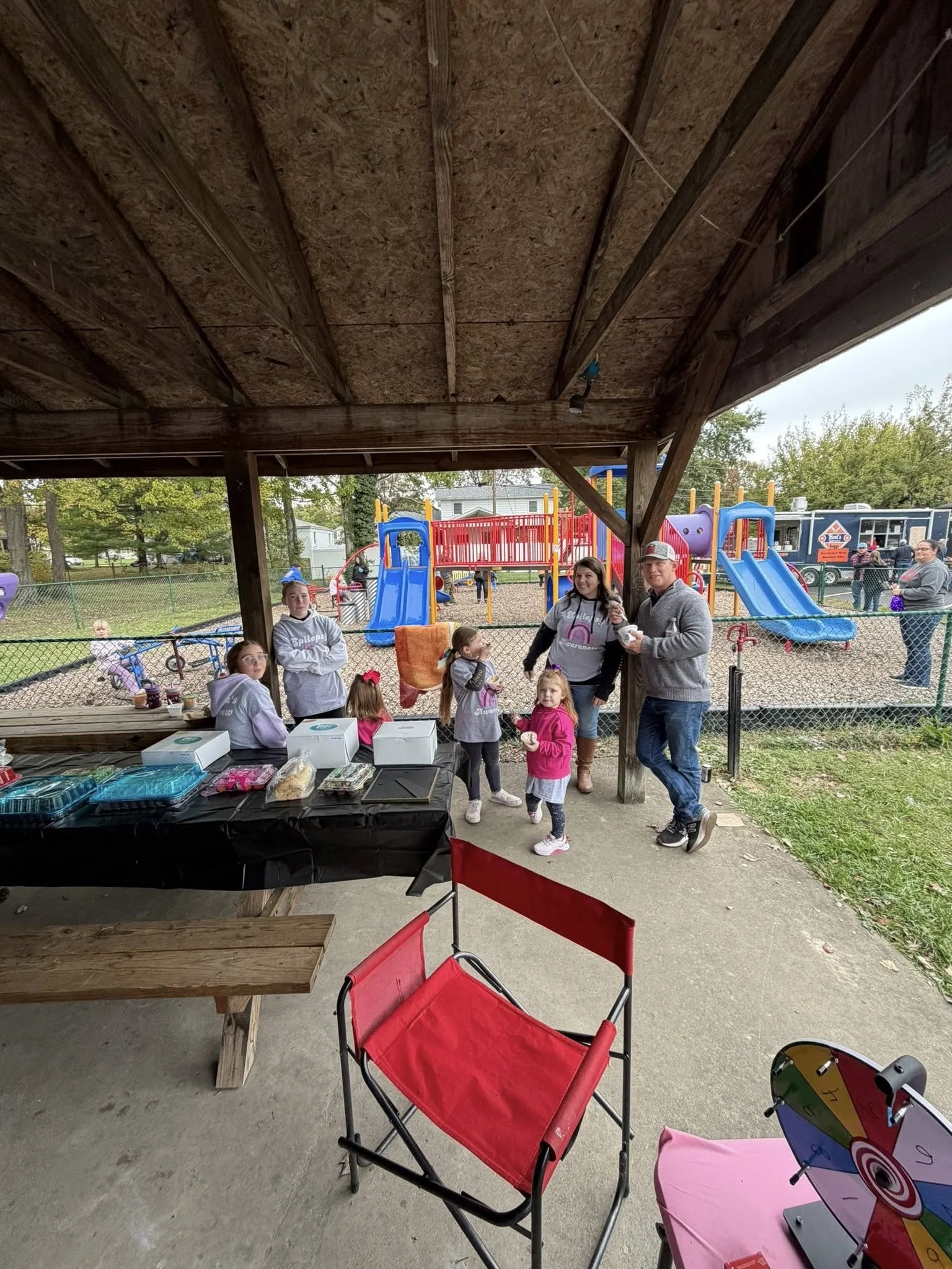 Children and adults gathering near a play area with slides, with a booth set up under a wooden shelter.