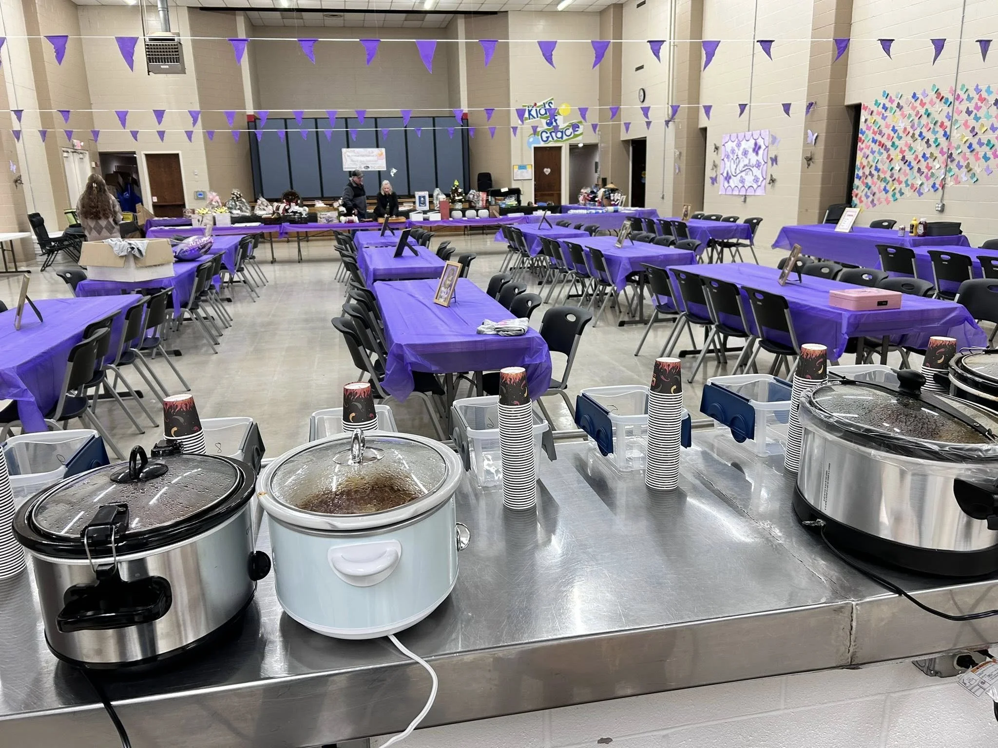 Party room decorated with purple triangular banners and tables covered with purple tablecloths. Beverage dispensers and cups are set up at the front, with a presentation area in the background. Kid's themed decorations are visible.