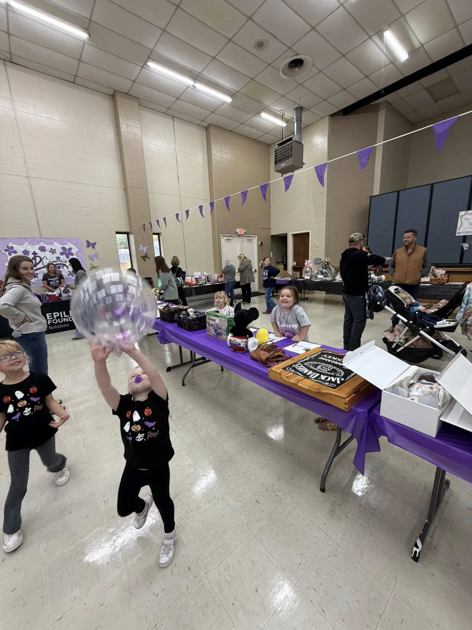 Children playing with a balloon at a community event in a large hall, tables with items and decorations, purple banners, and people browsing