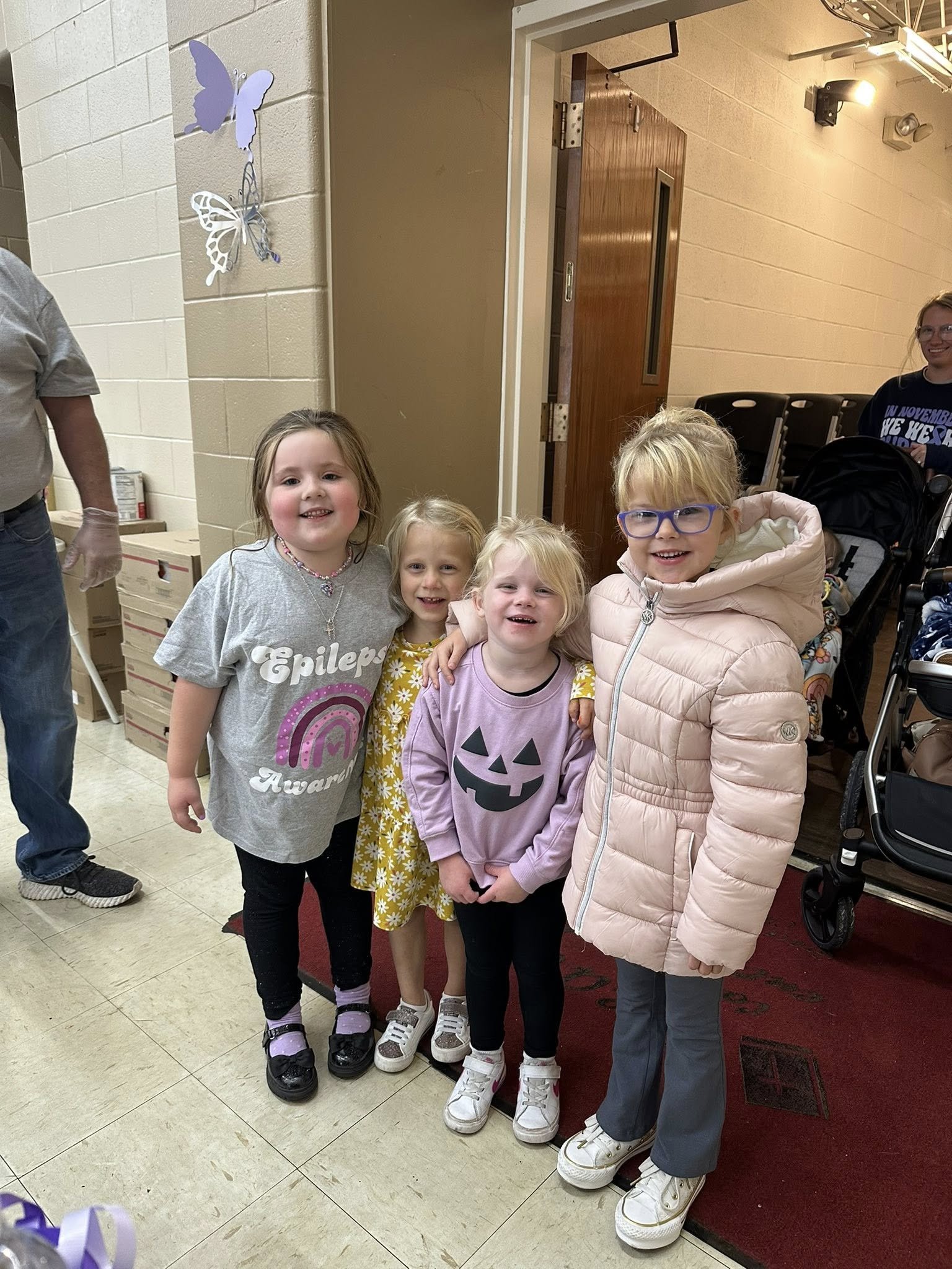 Four young girls smiling and standing together indoors, with colorful butterfly decorations on the wall in the background.