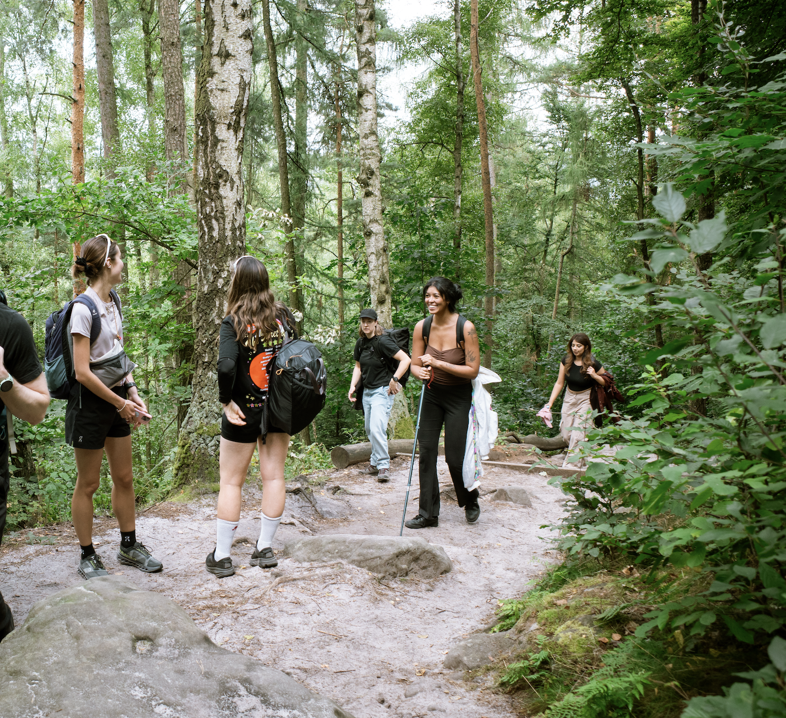 A group of young women hike on a forest trail surrounded by tall trees and green foliage.