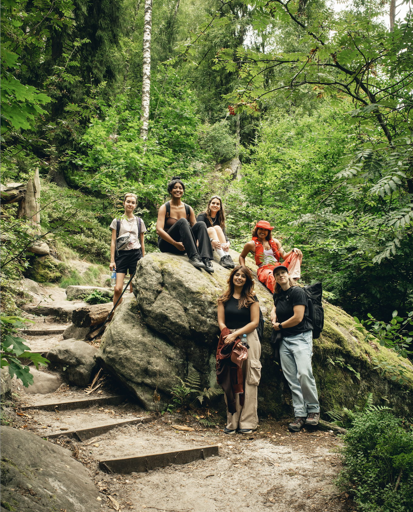 Group of six women hiking and resting on a large rock in a lush green forest.