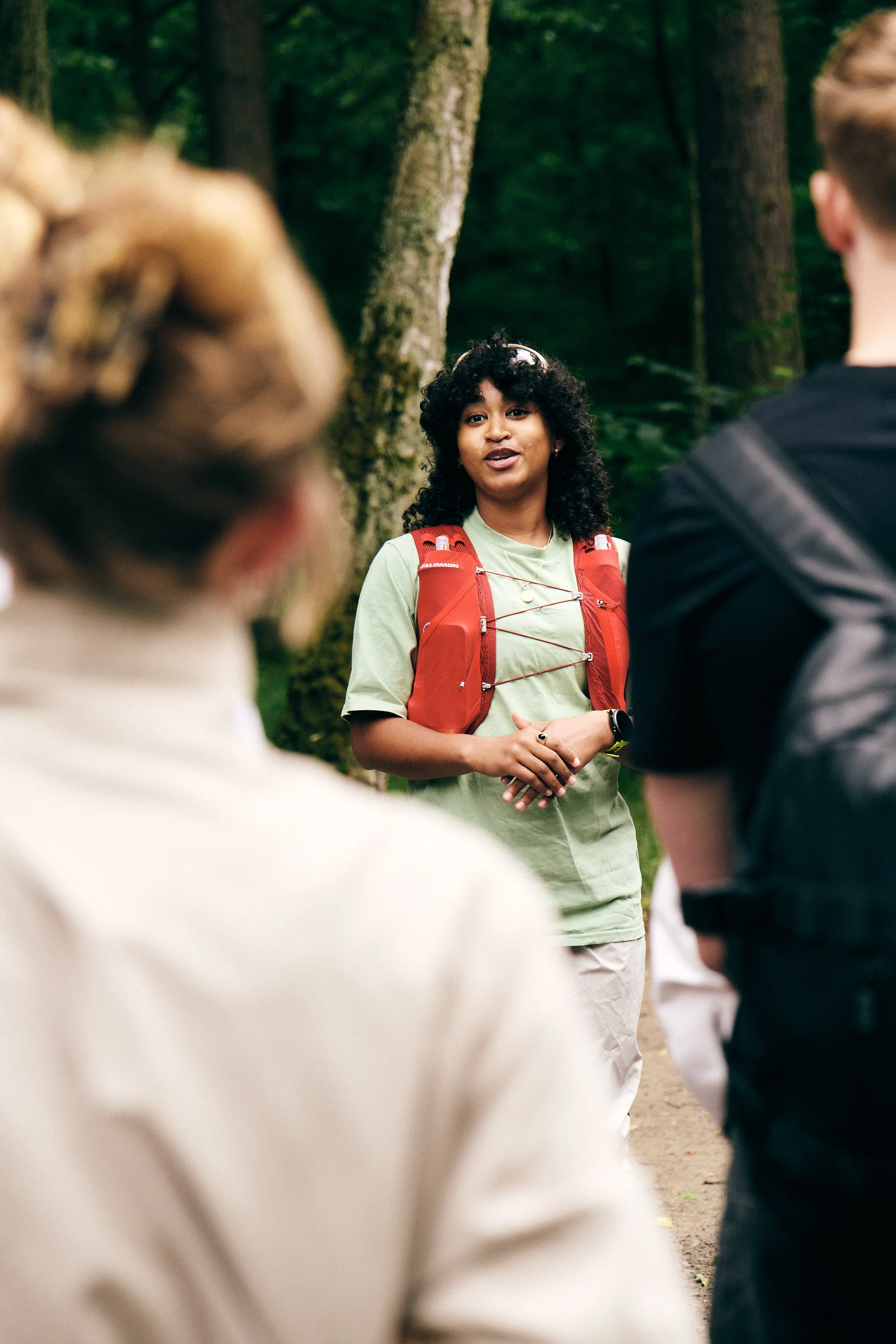 A woman with curly black hair, wearing a green shirt and a red backpack, speaks to a group of people in a forest.