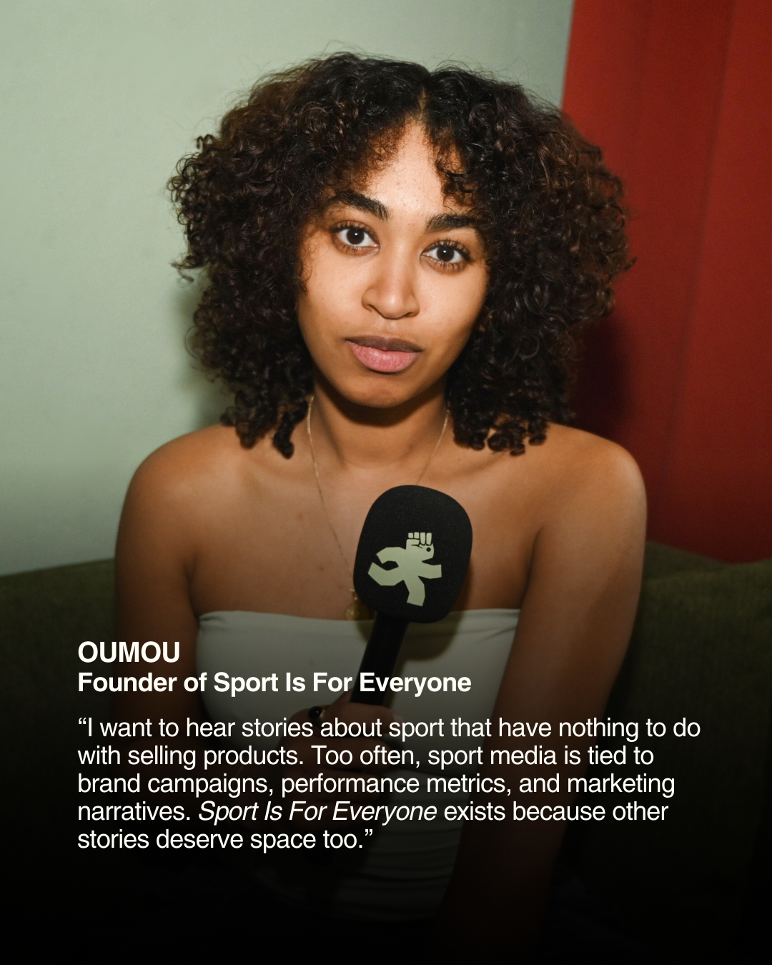 A young woman with curly hair holding a microphone with a sports activism logo, sitting on a green couch in front of a light wall with a red panel, speaking about sports stories.
