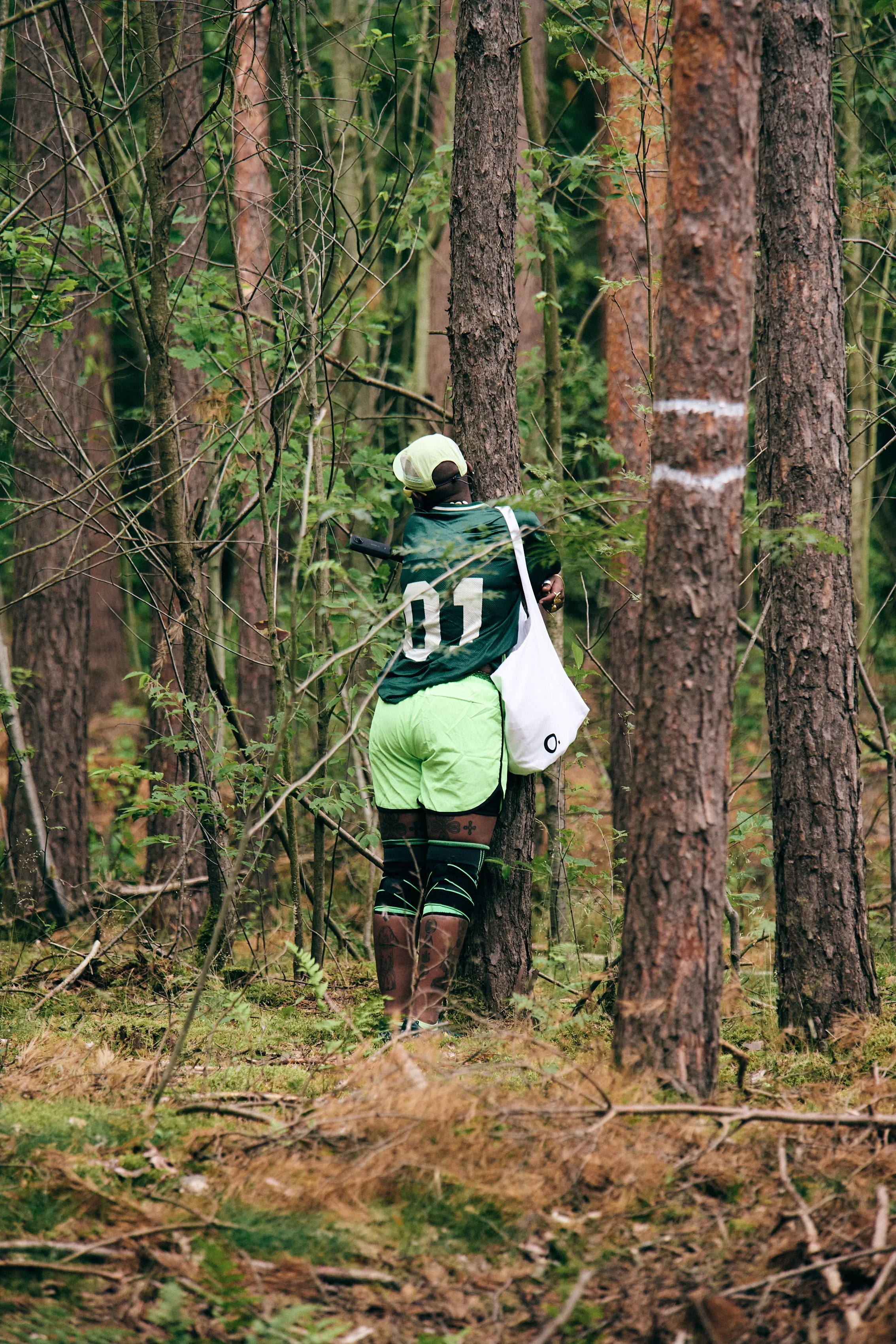 Person standing in a dense forest, wearing a green sports outfit, black knee-high socks, a white cap, and carrying a white bag.