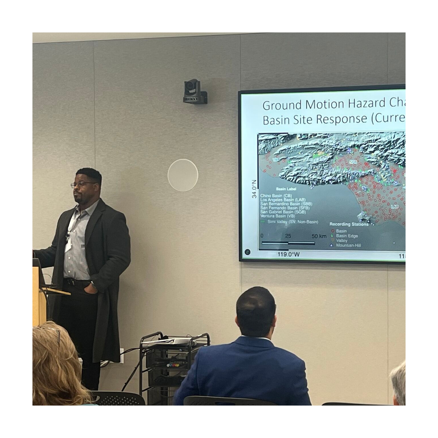 A presenter giving a technical talk in front of a projected slide titled “Ground Motion Hazard Characterization,” with attendees seated and watching.