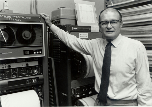 Black-and-white photo of a Professor Bolt standing beside seismic recording equipment labeled “U.S.G.S.–A.S.L.,” symbolizing early earthquake monitoring and data collection technology.