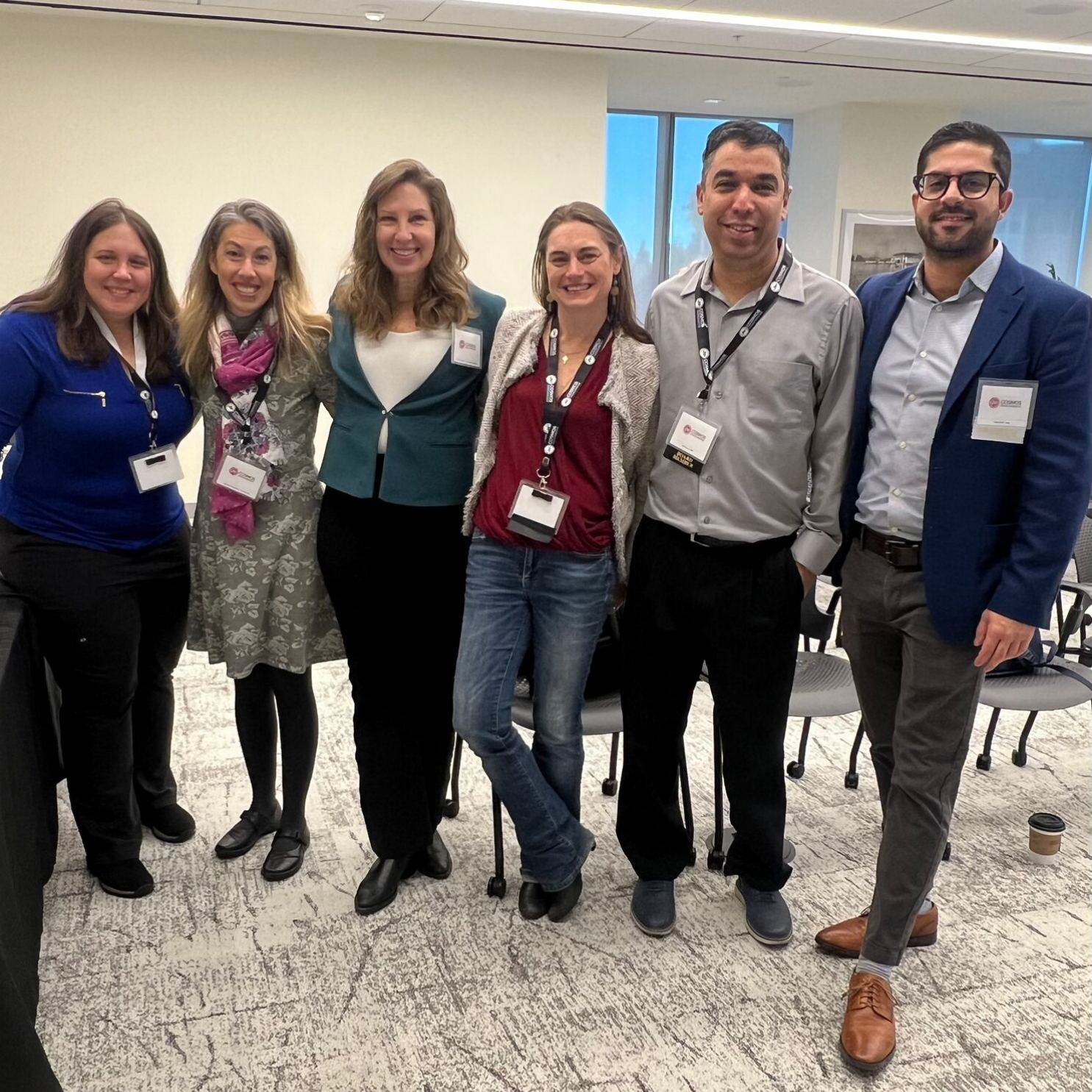 A group of six conference attendees smiling for a photo at a professional event, standing in a meeting room with badges and name tags