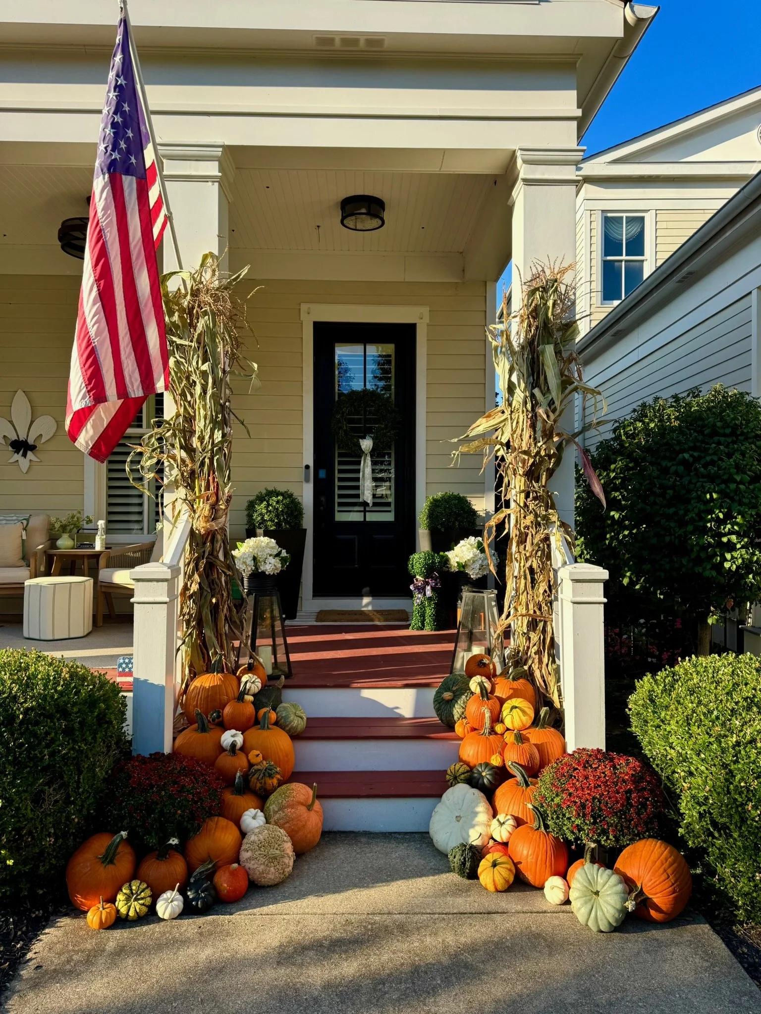Front porch decorated for fall with pumpkins, gourds, and flowers, American flag, corn stalks, and chairs.