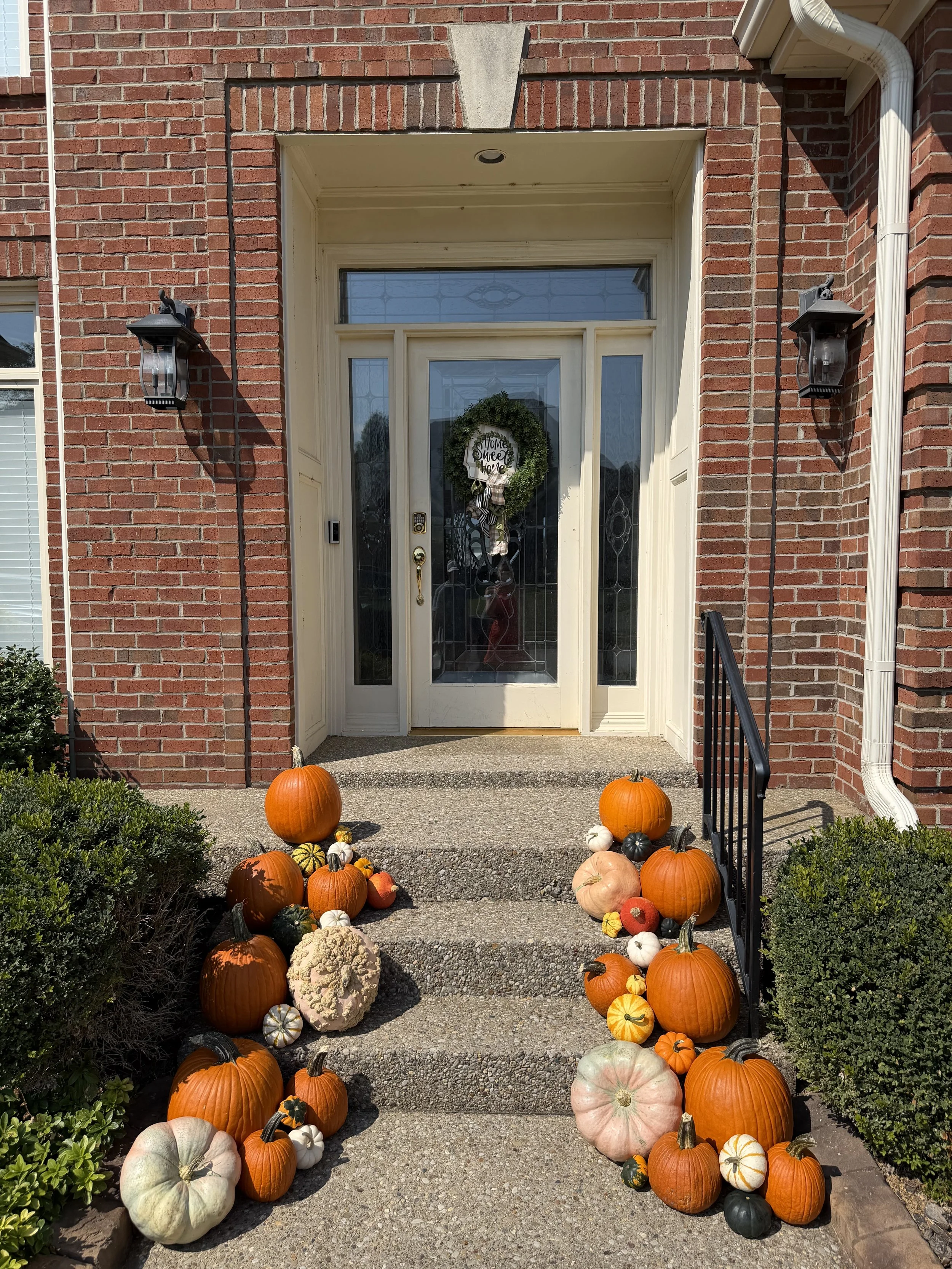 House entrance decorated with an assortment of pumpkins, gourds, and squash on the front steps, with a wreath hanging on the door that says 'Home Sweet Home'.