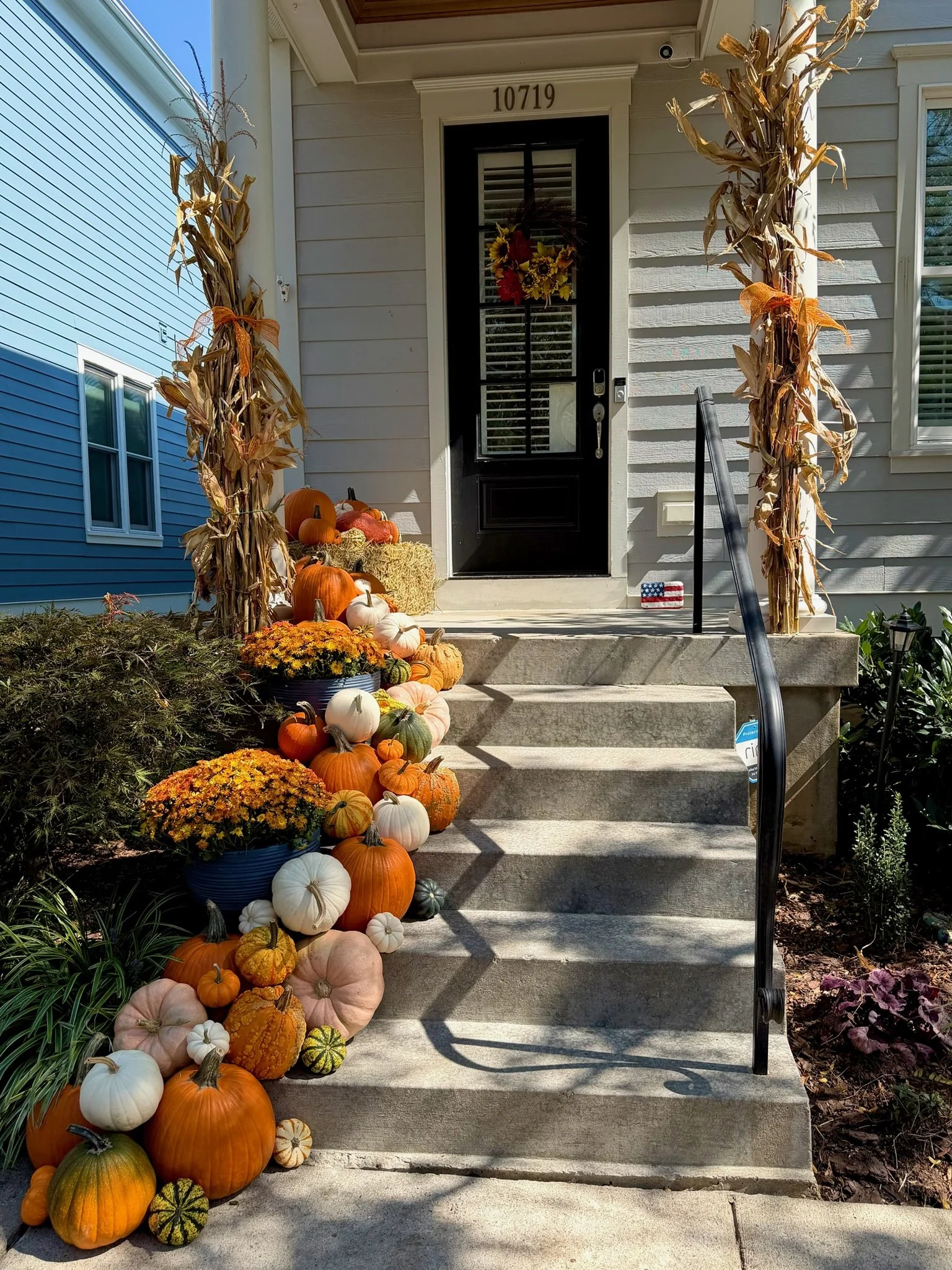 Front porch decorated for fall with an array of pumpkins and gourds on the stairs and yard, corn stalks on either side of the door, and a small American flag decoration by the door.