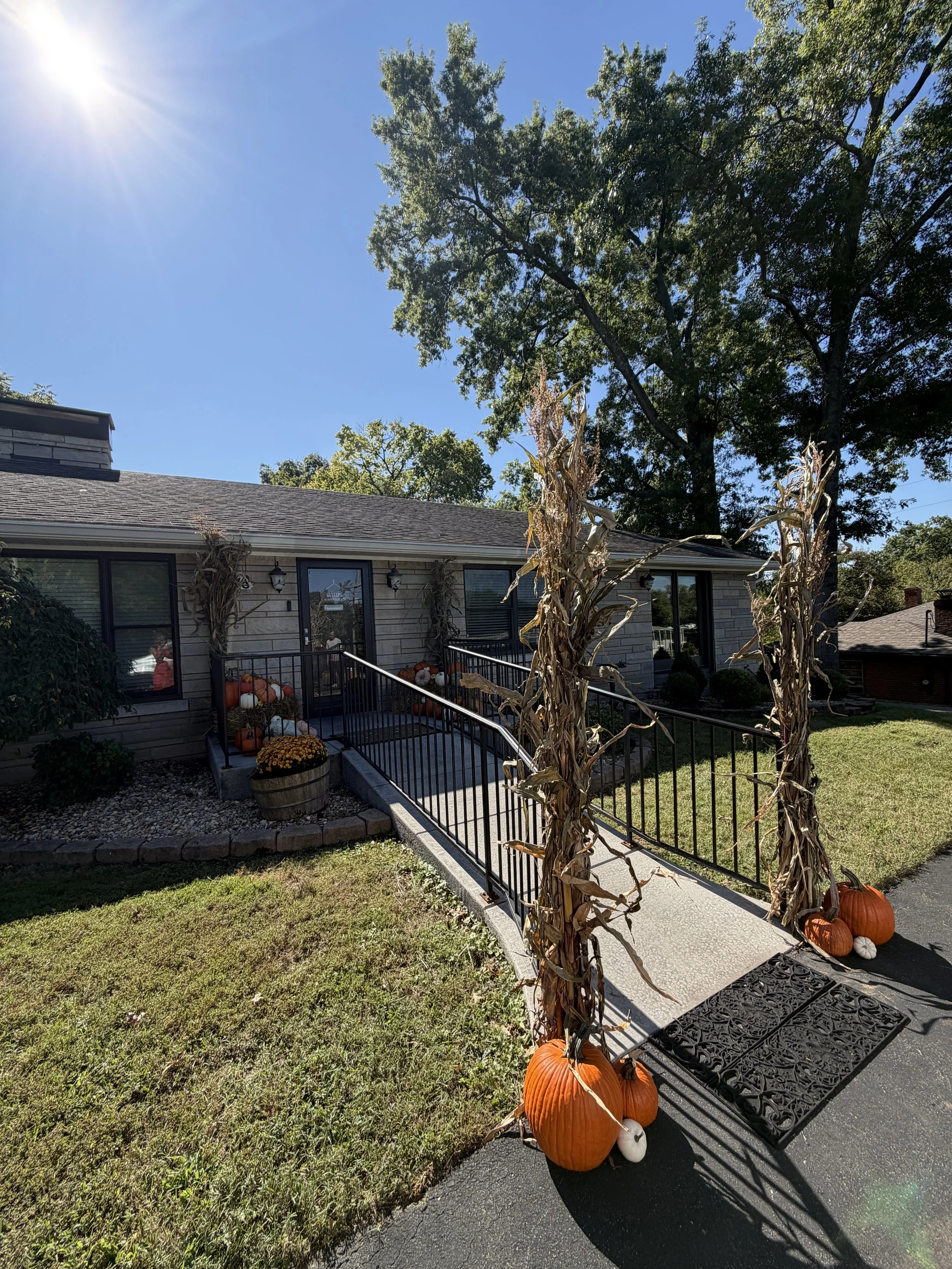 A house decorated with pumpkins, gourds, and dried cornstalks for fall or Halloween, with a front porch, black metal railing, and a pathway leading to the entrance, surrounded by a well-kept lawn and tall trees under a clear blue sky.