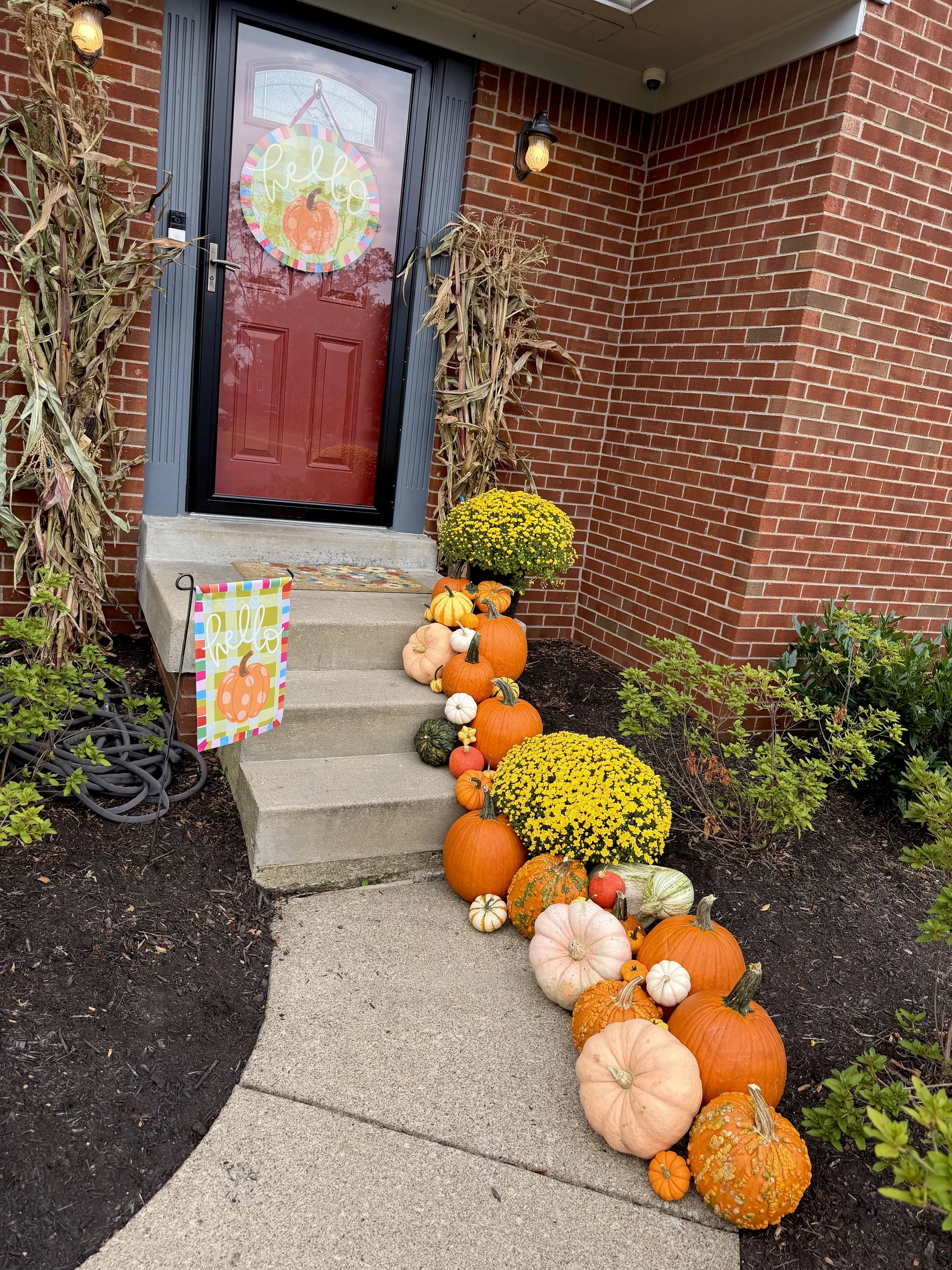 Front porch entrance decorated with fall and Halloween decorations, including a group of pumpkins, gourds, yellow chrysanthemums, a welcome sign with a pumpkin, and dried corn stalks.