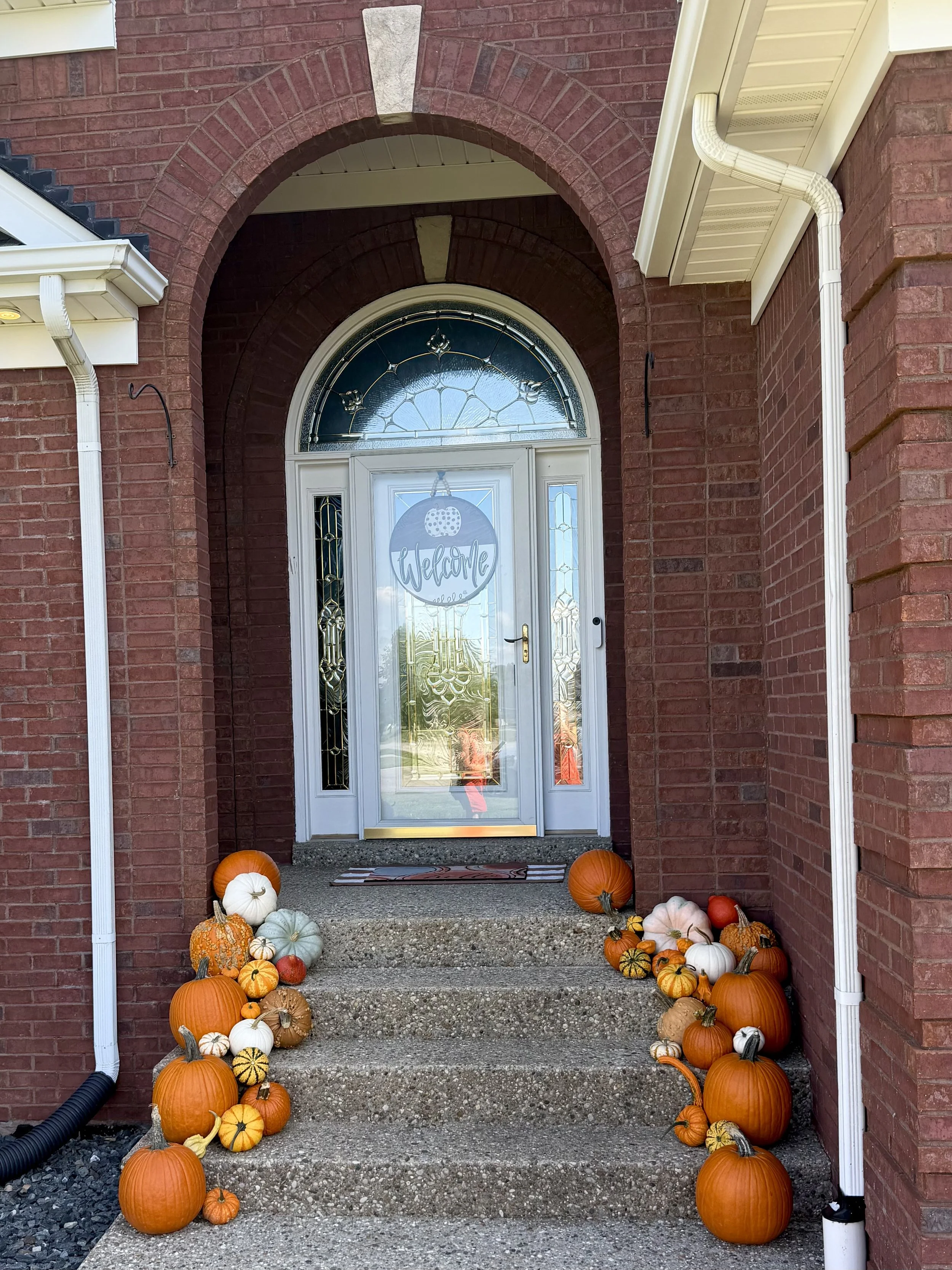 Front porch of a house with red brick walls, white door, and an archway, decorated with pumpkins of various sizes and colors, with a "Welcome" sign hanging on the door.
