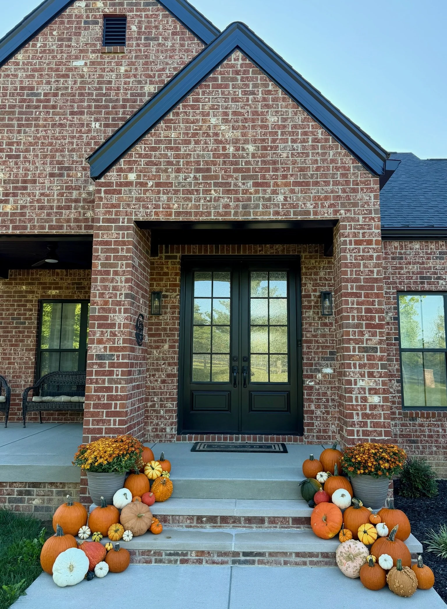 Fall decorations on front porch with pumpkins and flowers, black front door, brick house, porch lights, and a doormat.