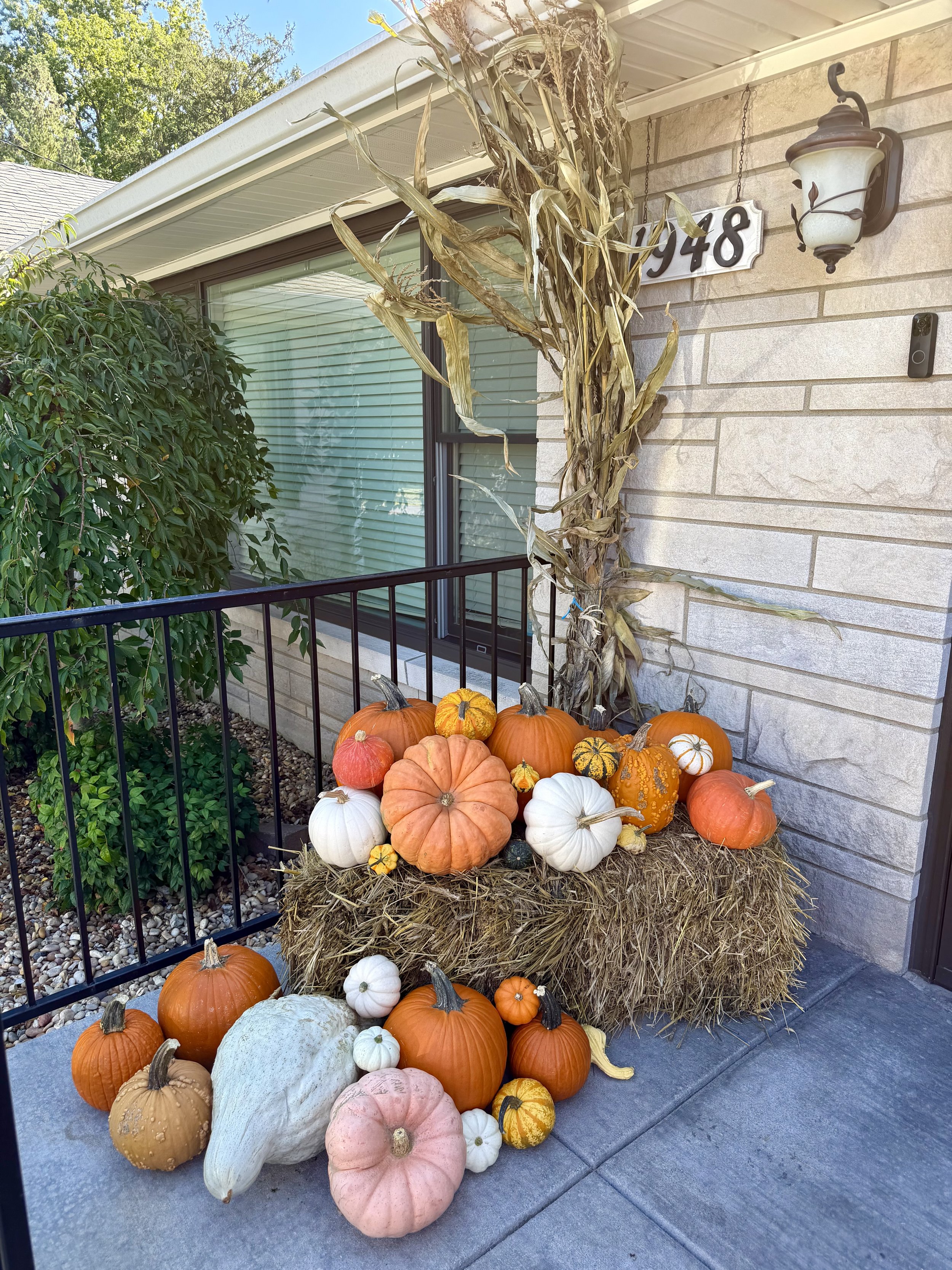 A window with closed blinds, a porch with a black metal railing, a bunch of gourds and pumpkins of various colors and sizes arranged on a hay bale, and a dried cornstalk plant next to the house wall.