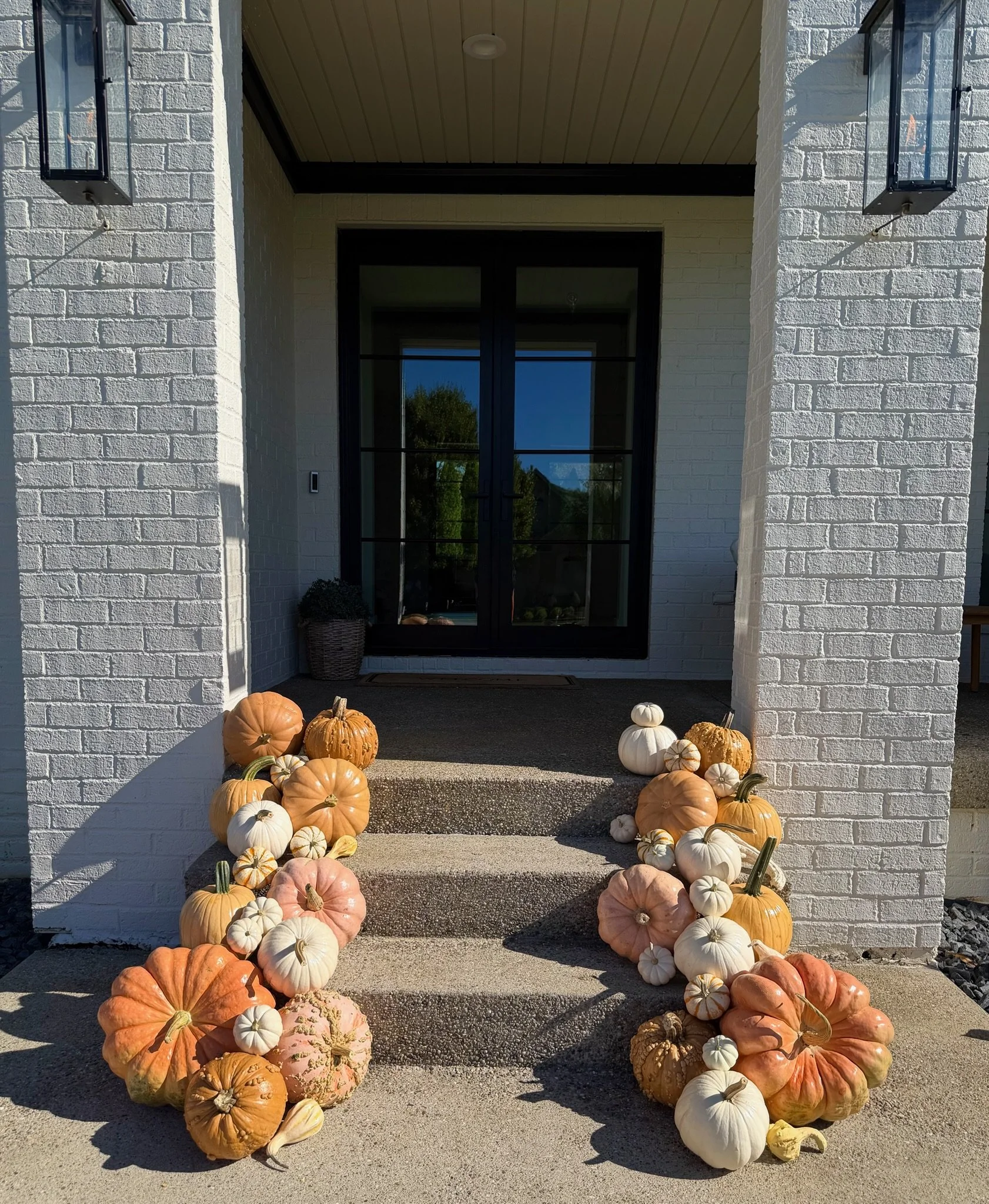 A front porch with white brick columns and a black door, decorated with various pumpkins and gourds in shades of orange, white, pink, and green, arranged along the steps and porch area. Pumpkin porch decorating Louisville KY
