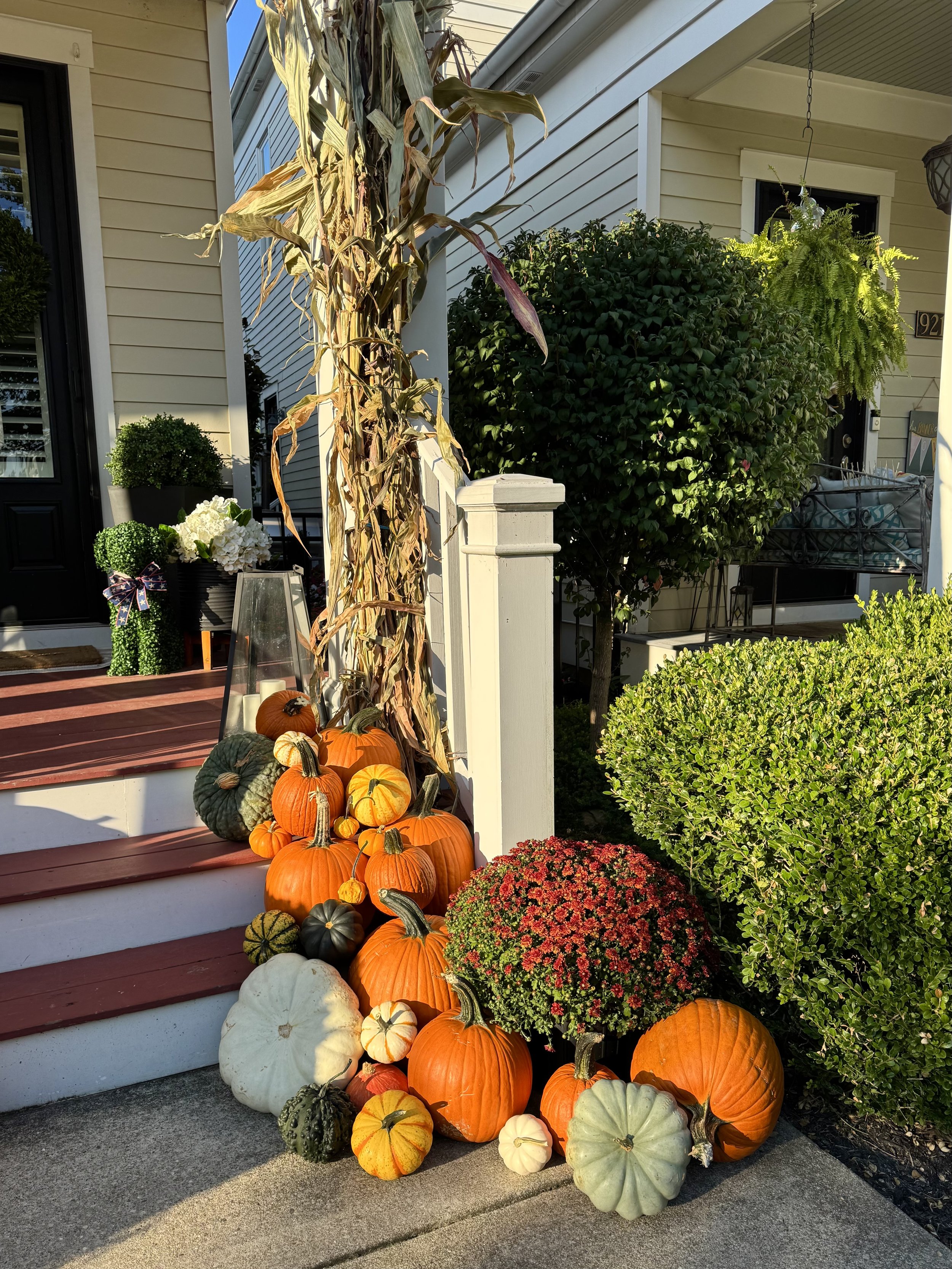 Pumpkins of various sizes and colors, including orange, green, white, and striped varieties, arranged at the base of a porch. Seasonal decorations with flowers and greenery are visible around the pumpkins, and the setting appears to be autumn.
