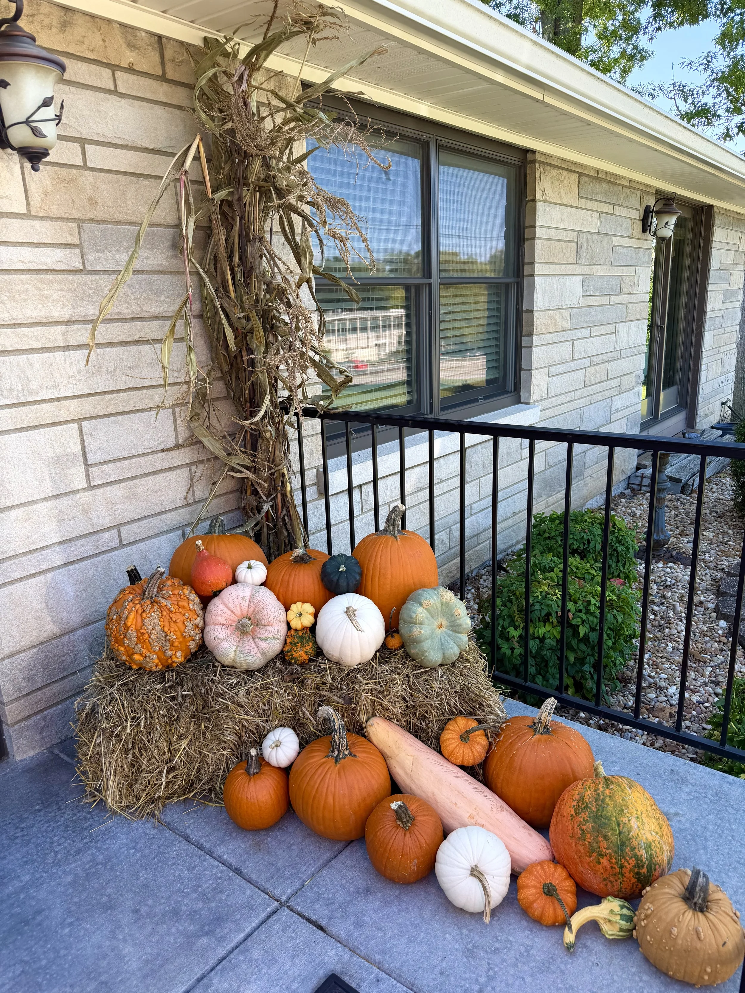 A collection of pumpkins, gourds, and dried decorative corn arranged on a bale of hay outside a house with brick siding and large windows. Dried corn stalks attach to the house wall, and outdoor lights are mounted beside the windows.