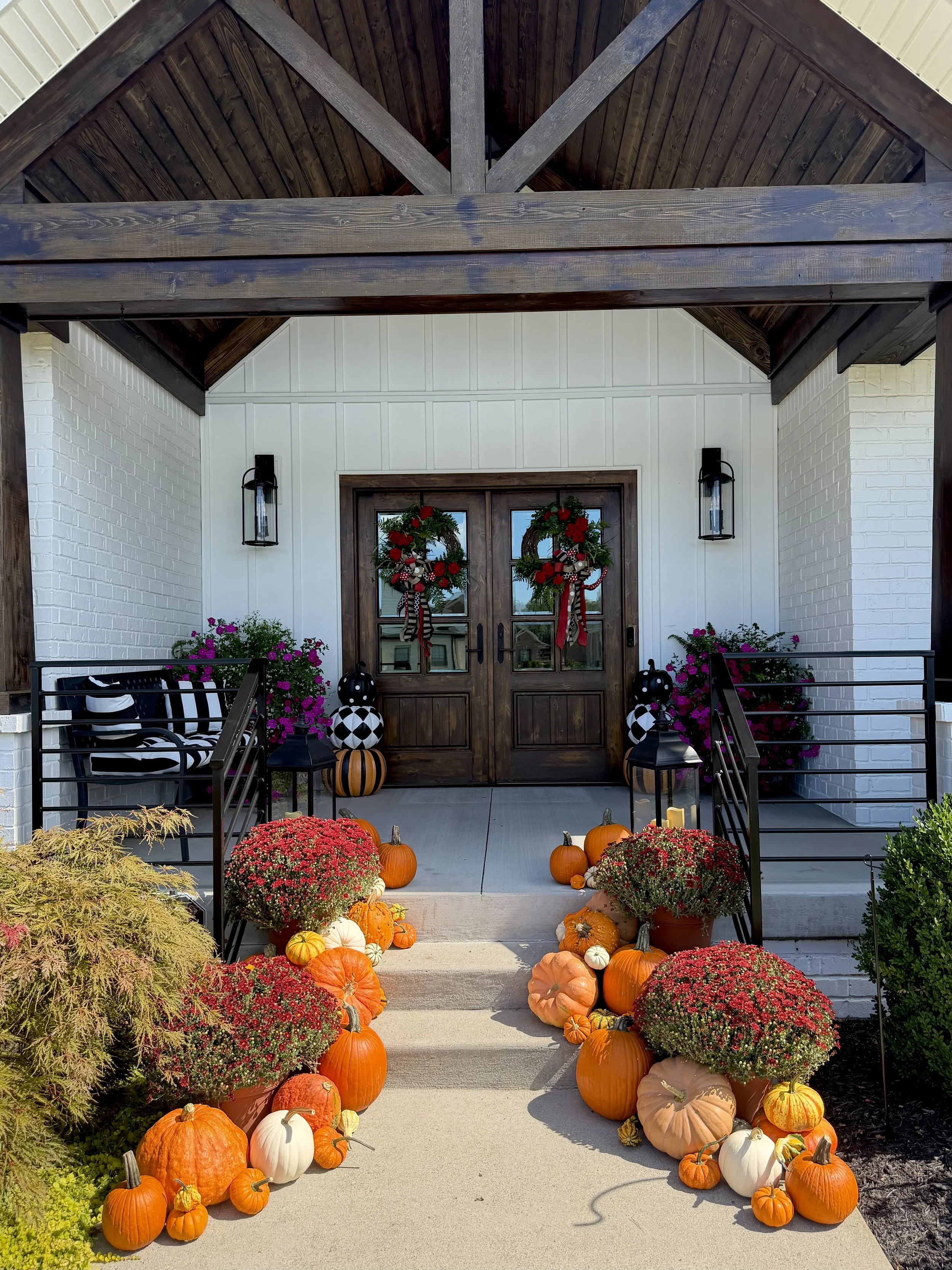 Front porch decorated for fall with pumpkins, gourds, and mums. Two wreaths hang on wooden double doors, flanked by black lanterns. Decorative pumpkins and fall flowers line the steps and porch area.