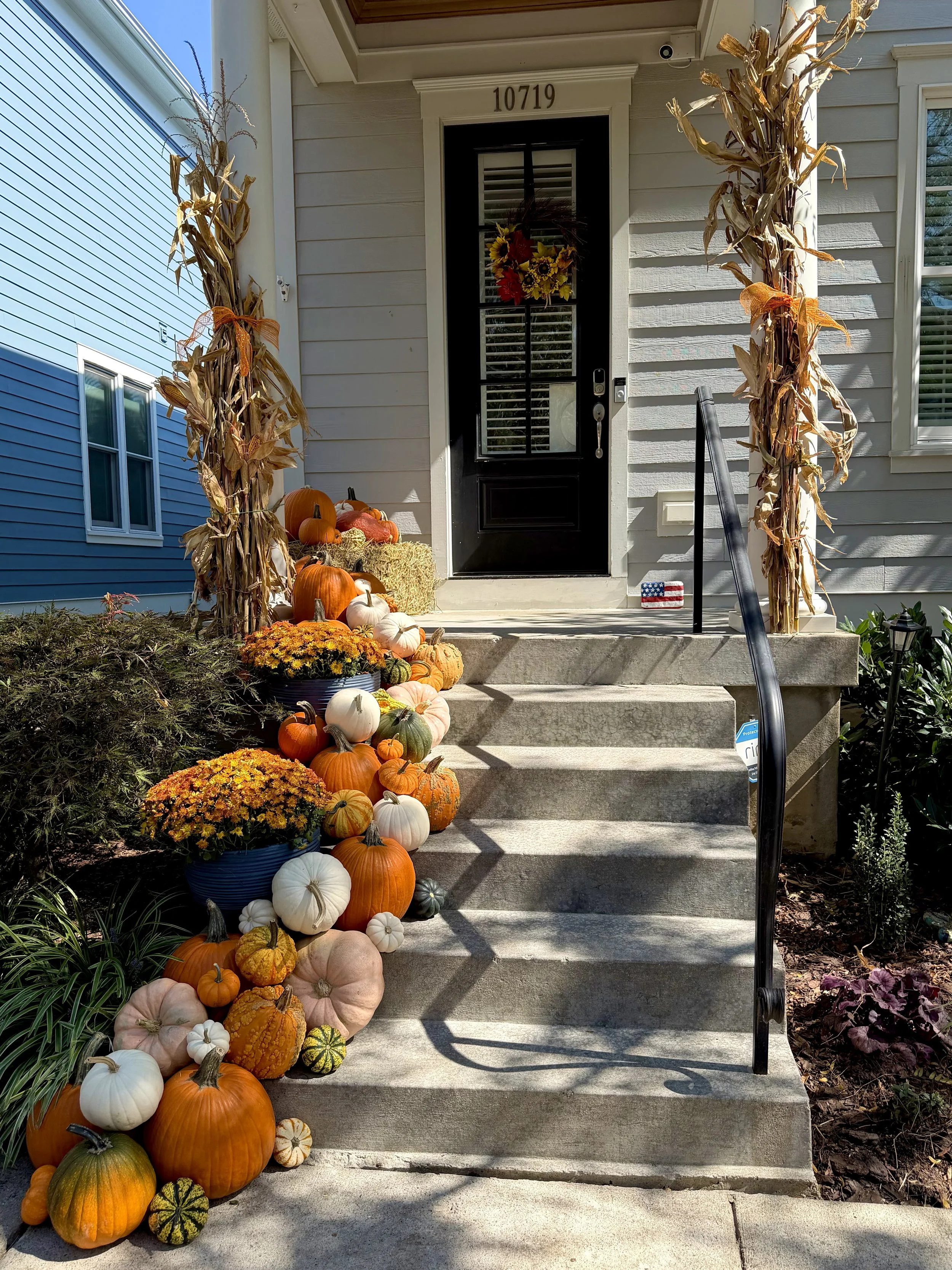 Decorated front porch with fall harvest pumpkins, gourds, and chrysanthemums on steps and on the side, two large corn stalks on either side of the door, and a wreath with sunflowers hanging on the black door with house number 10719.