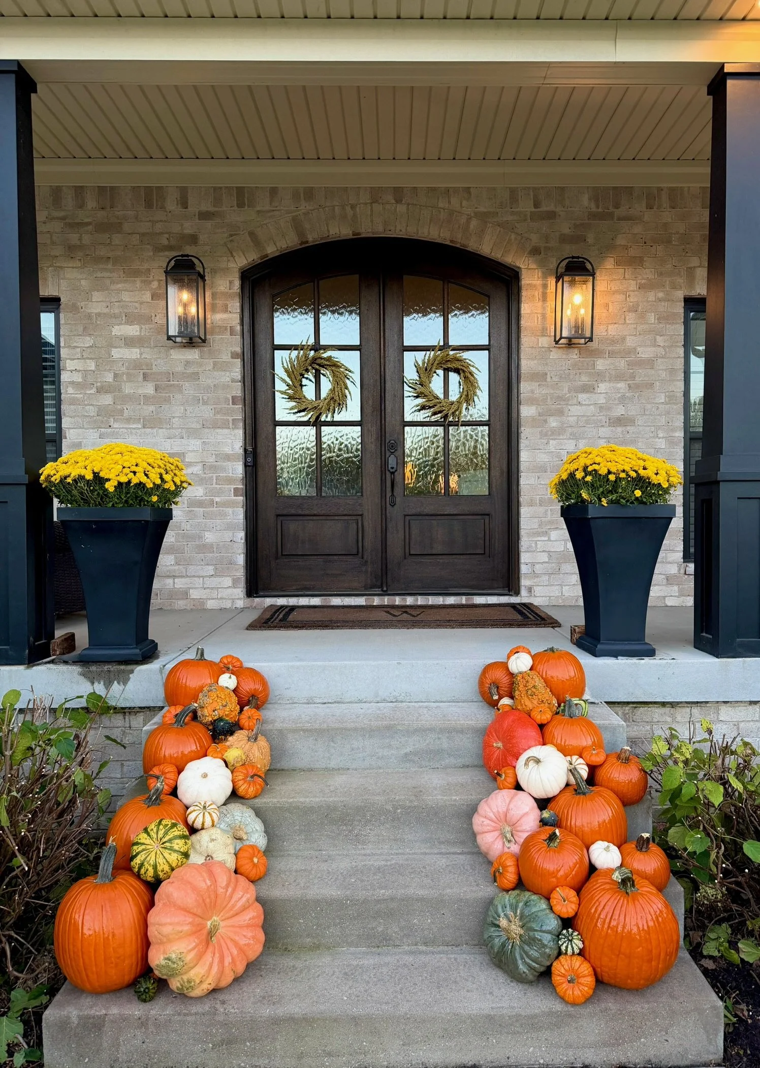 Front porch decorated for fall with  pumpkins and gourds, yellow mums, wreaths on the door, and lantern-style lights.