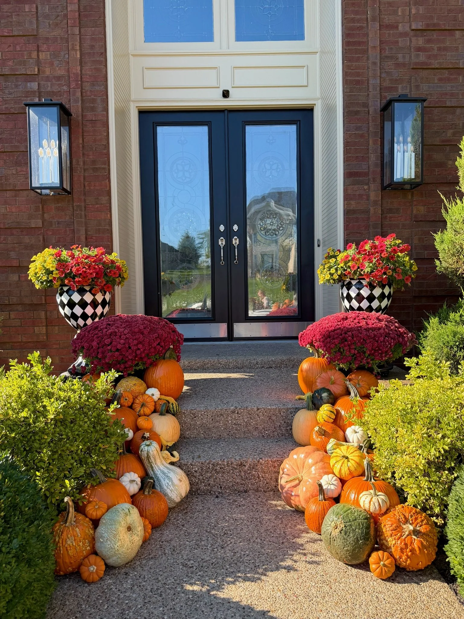 Front entrance of a brick house decorated for fall with pumpkins, gourds, and potted mums flanking the doorway, and lanterns mounted on the brick wall.