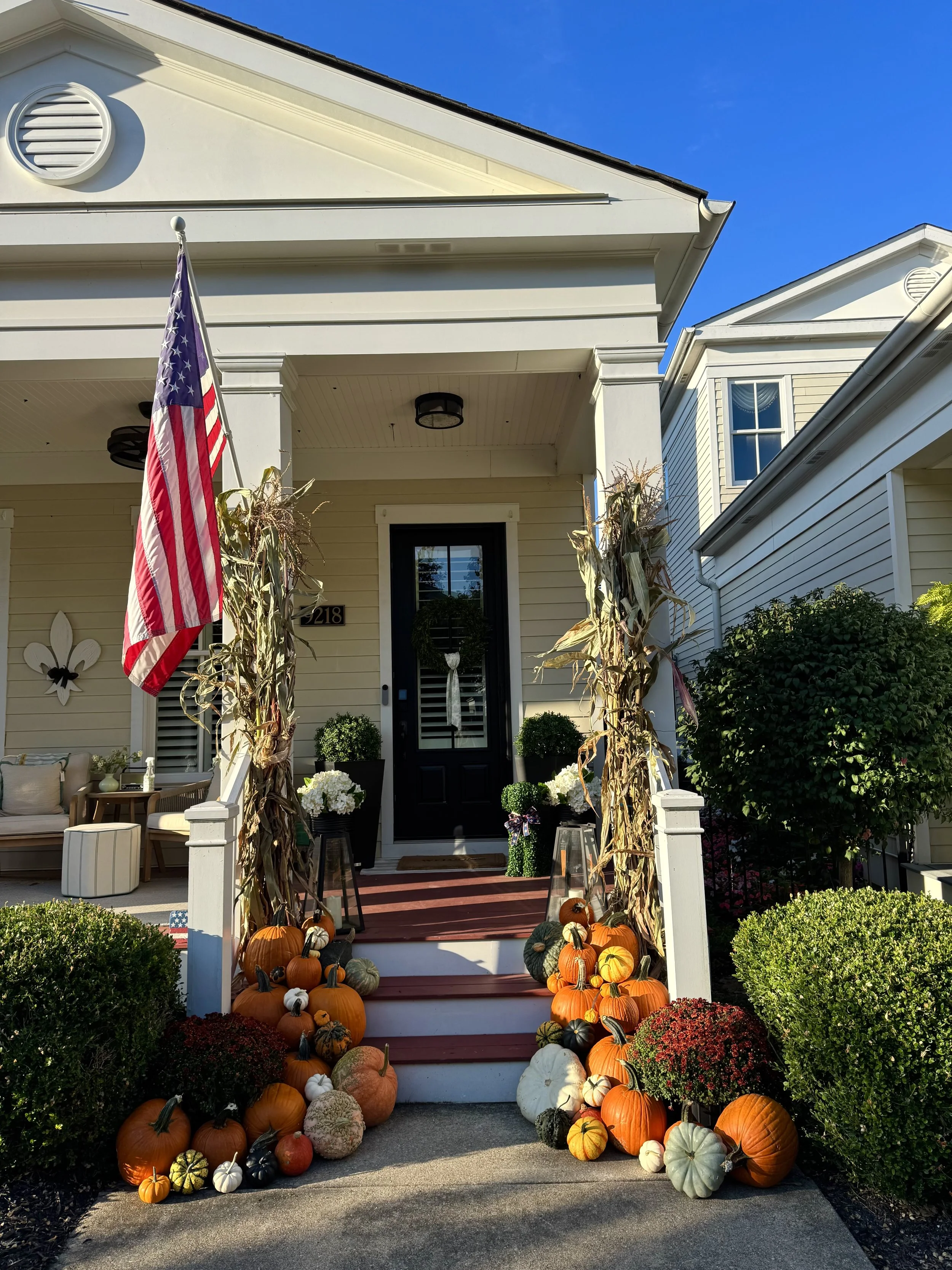 Front porch decorated with pumpkins and mums for fall, with an American flag on a porch post and a wreath on the door, under a sunny blue sky.