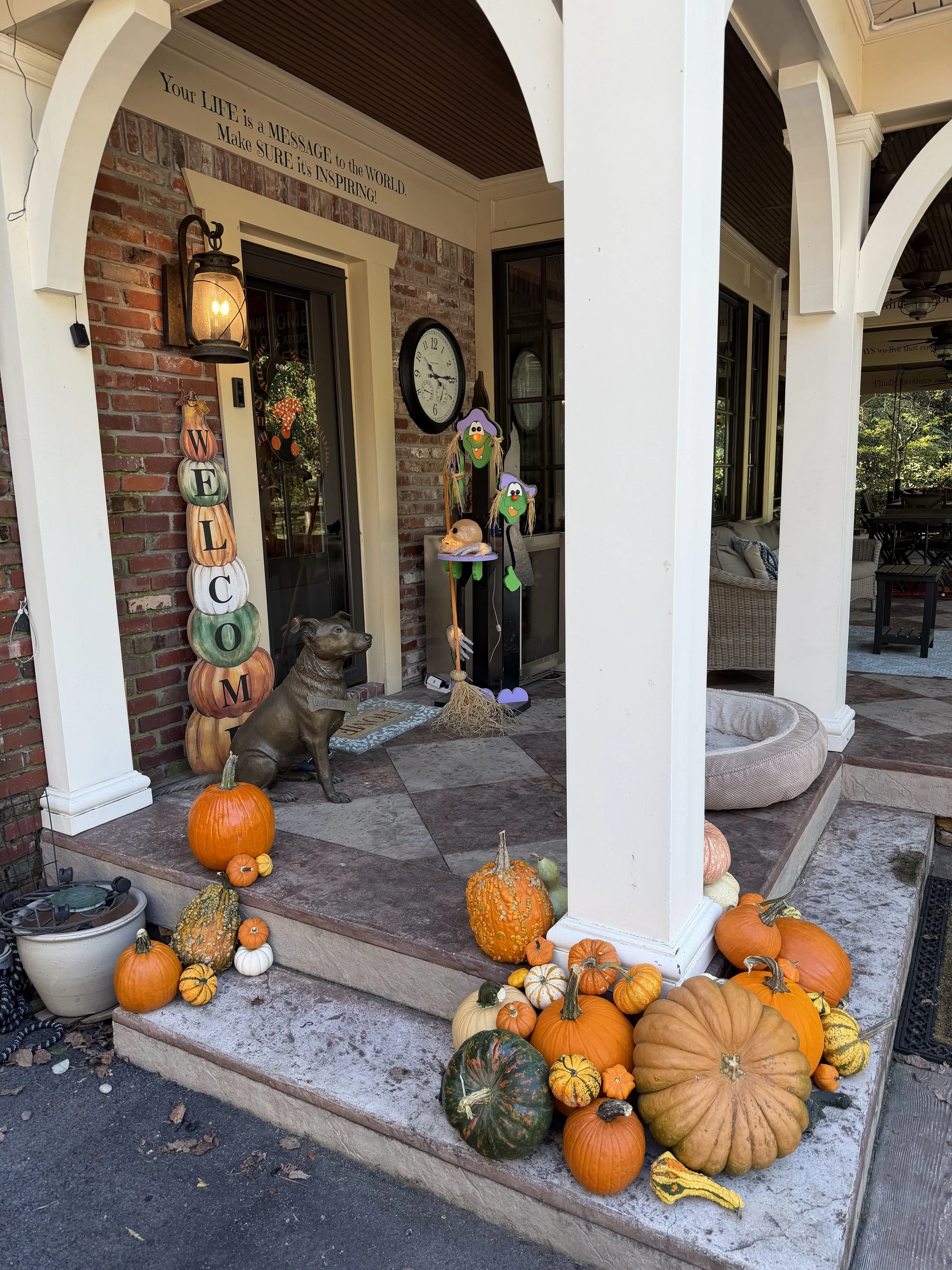 Front porch decorated with fall pumpkins and gourds, a weathered dog statue, a Welcome sign, Halloween masks, and seasonal decor, with a brick wall and windows on the house.