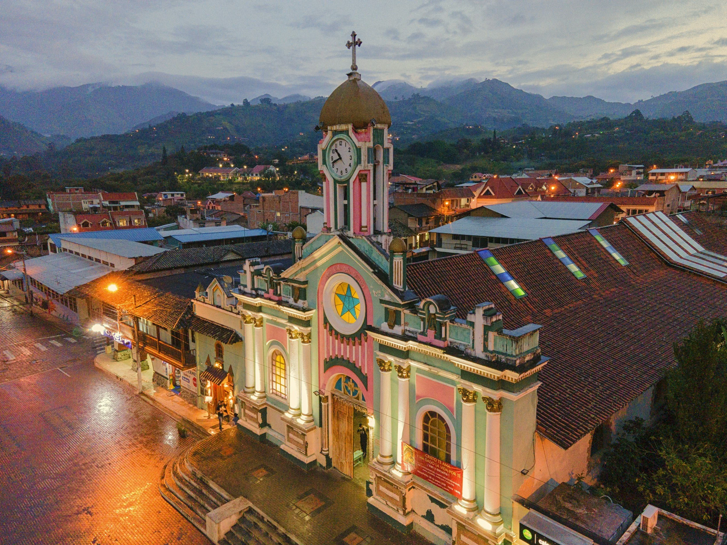 A colorful church with a pink and white facade, a green clock tower, and a cross on top, situated in a town at dusk surrounded by hilly terrain, with streetlights illuminating the cobblestone streets.