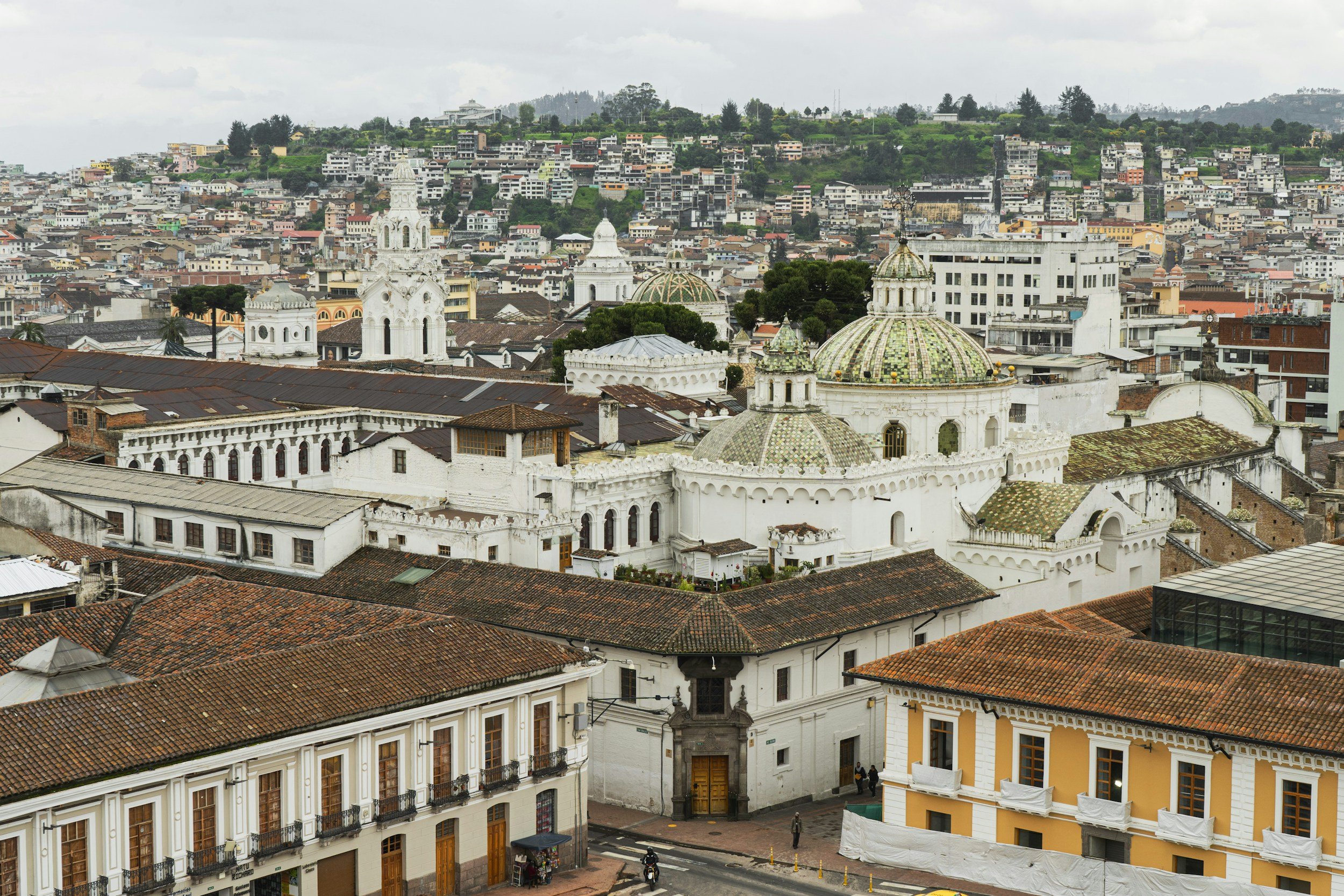 Cityscape of historic downtown, featuring colonial-style white buildings with red tile roofs, a church with domed towers, and a hilly area with dense residential housing and greenery in the background.