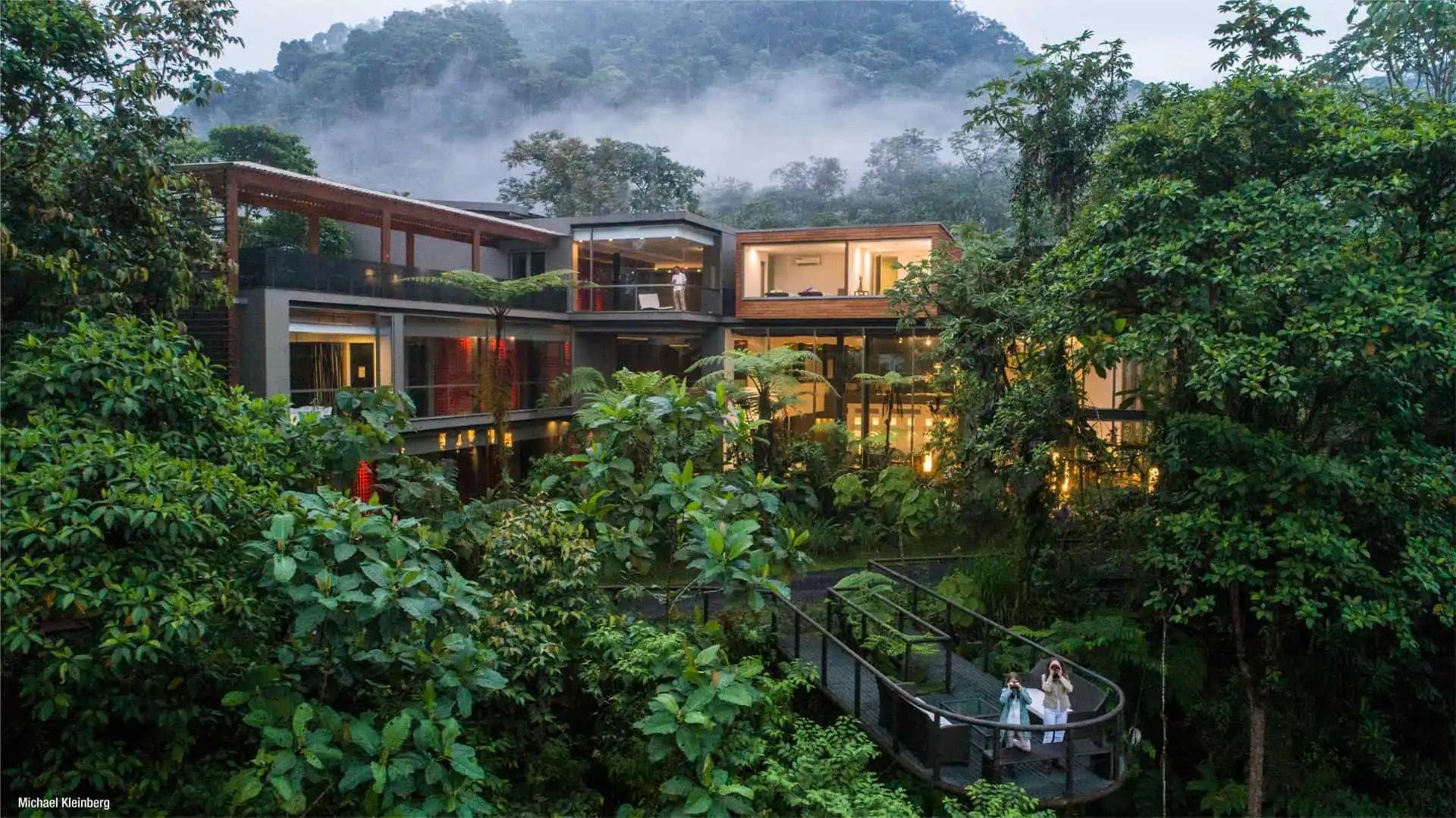 Modern multi-story house with large glass windows and balconies surrounded by lush green trees and plants, with misty mountains in the background.