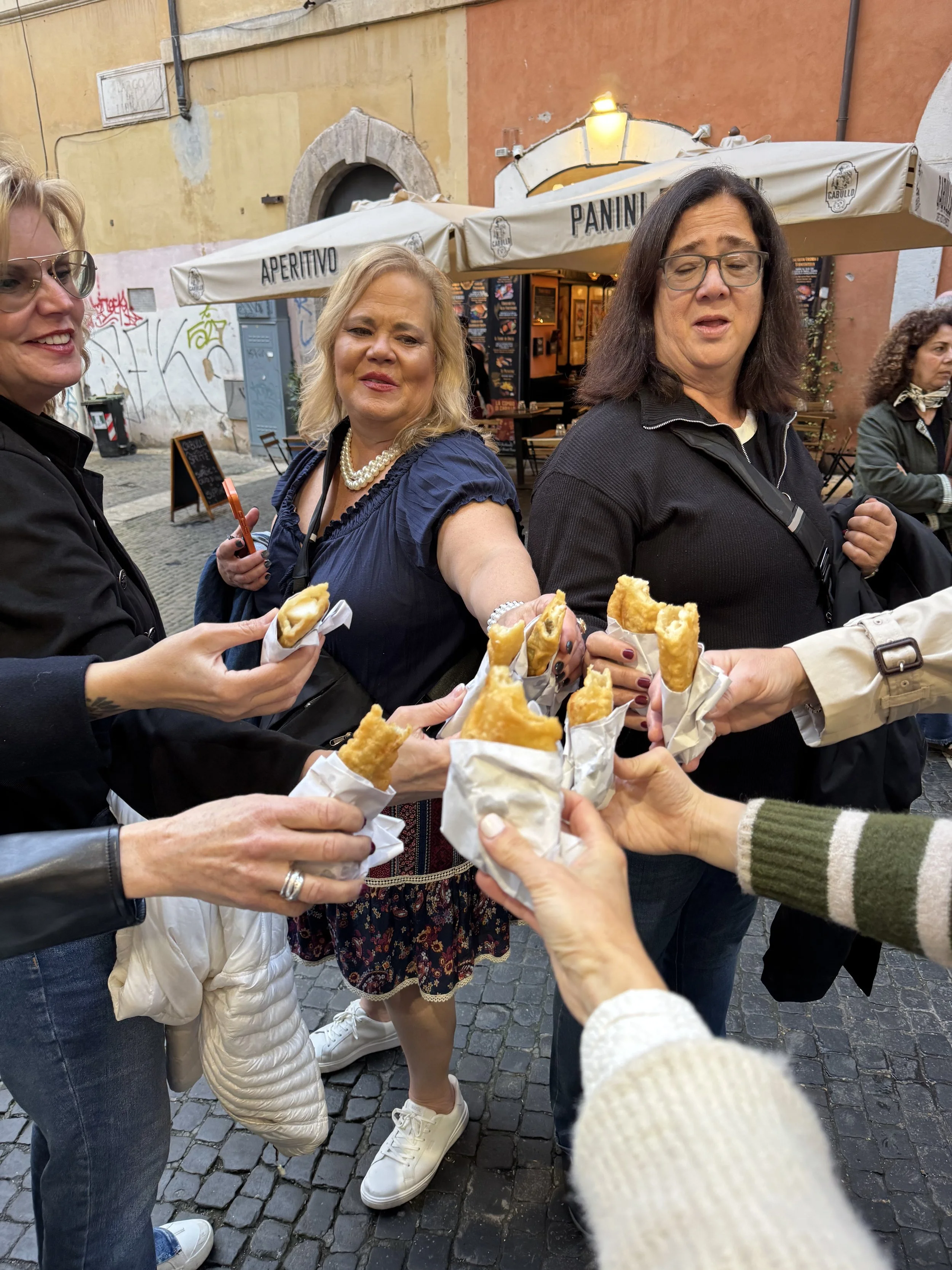 People gathered outdoors, holding fried food in wrappers, sharing a meal on a cobblestone street near a restaurant with outdoor seating and colorful buildings.