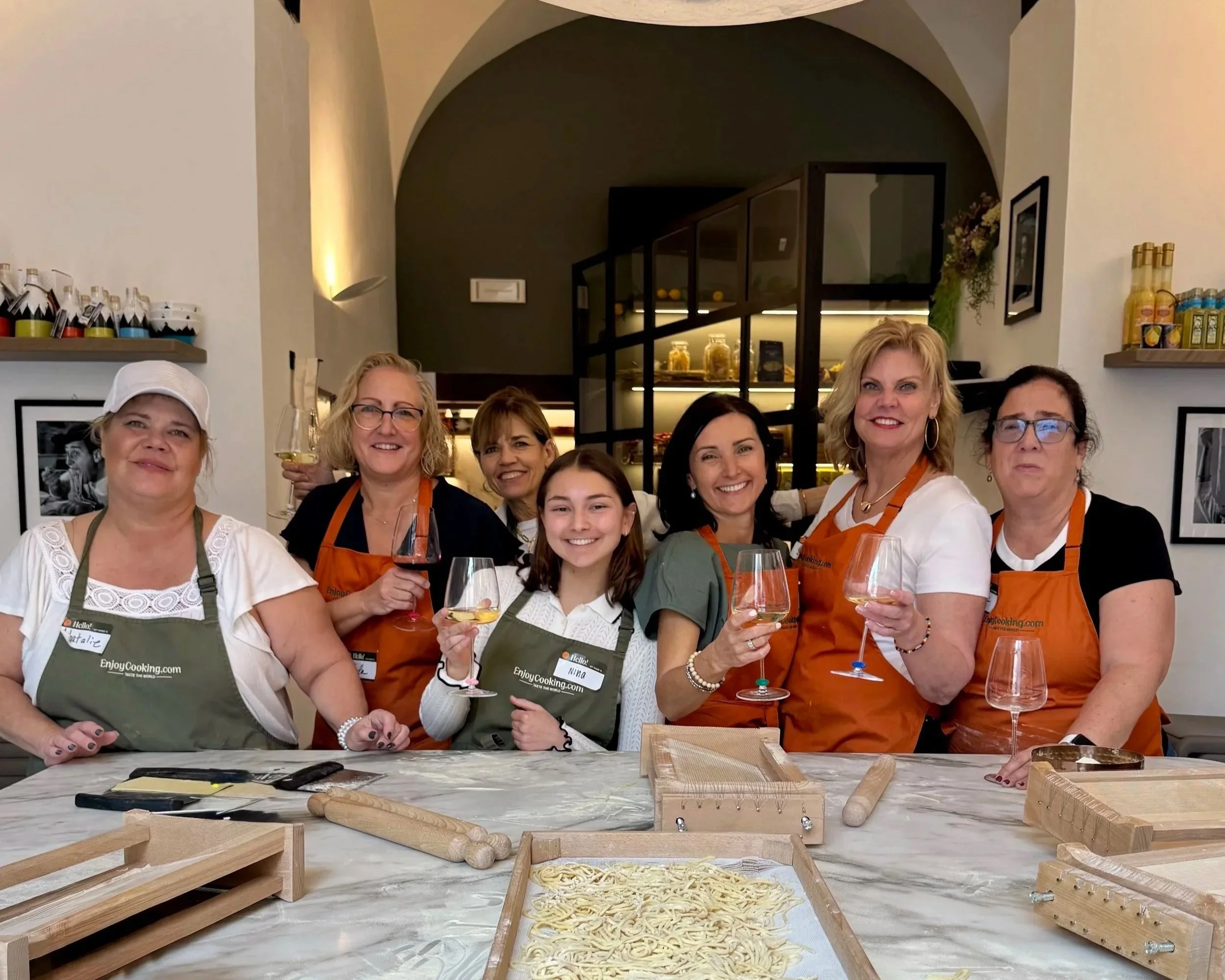 Group of women and girls gathered around a table in a cooking class, holding wine glasses, with pasta-making tools and fresh pasta in front of them, inside a modern kitchen setting.