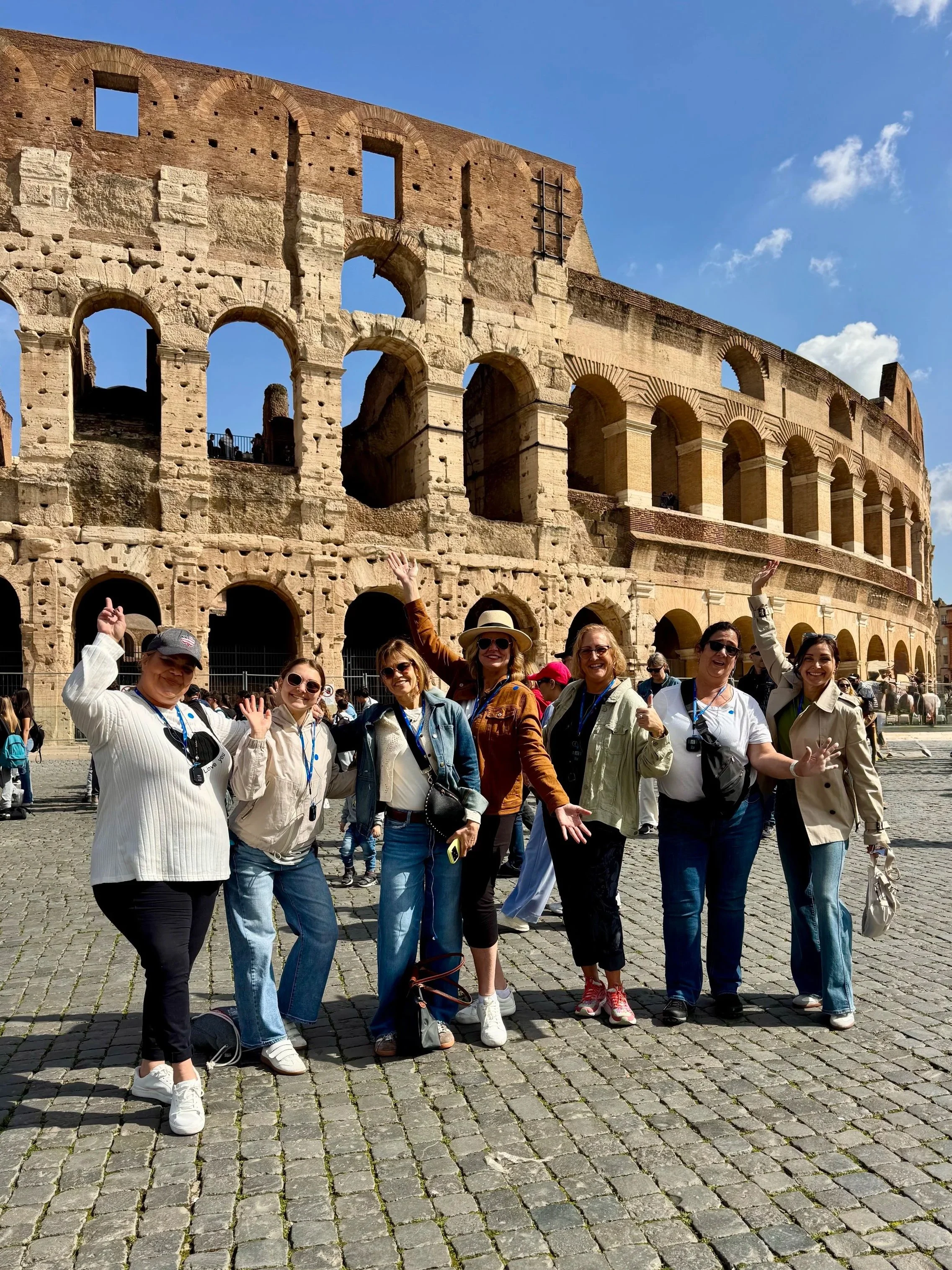 Group of tourists posing in front of the ancient Roman Colosseum in Rome, Italy, with clear blue sky in the background.