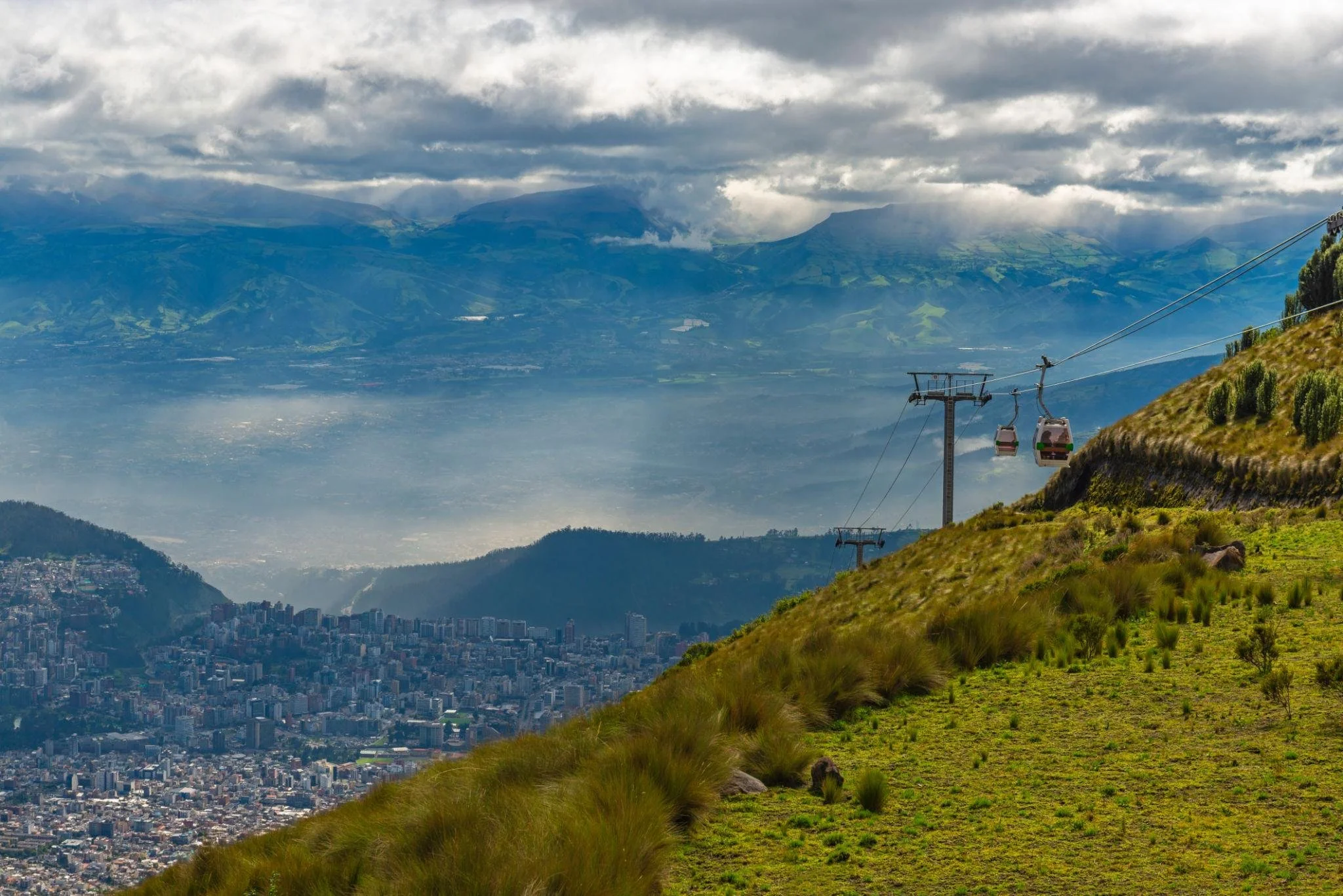 A mountain landscape with a city below and a cable car ascending the hillside surrounded by green vegetation, with mountains and clouds in the background.
