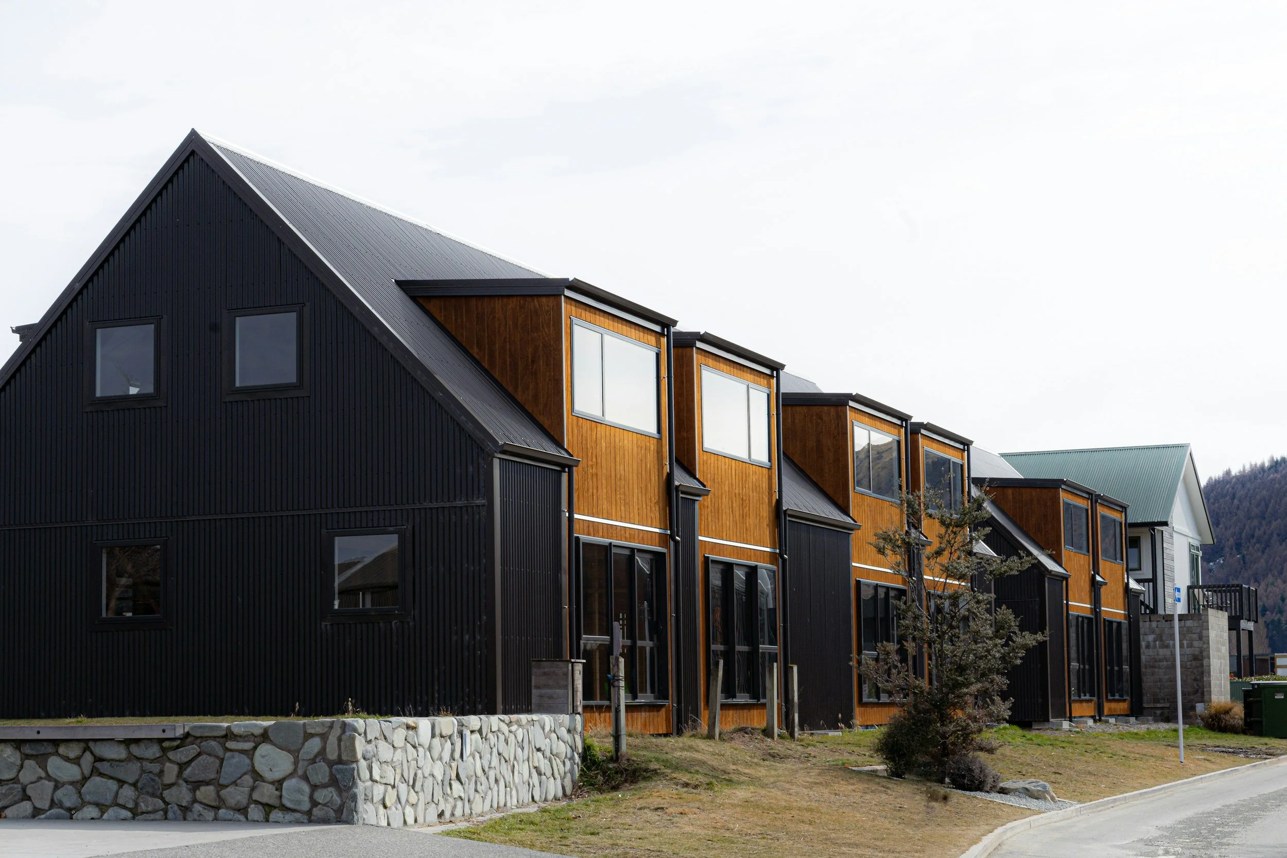 Modern multi-story residential buildings with black and wood siding, large windows, and a stone retaining wall, set in a suburban neighborhood with hills in the background.