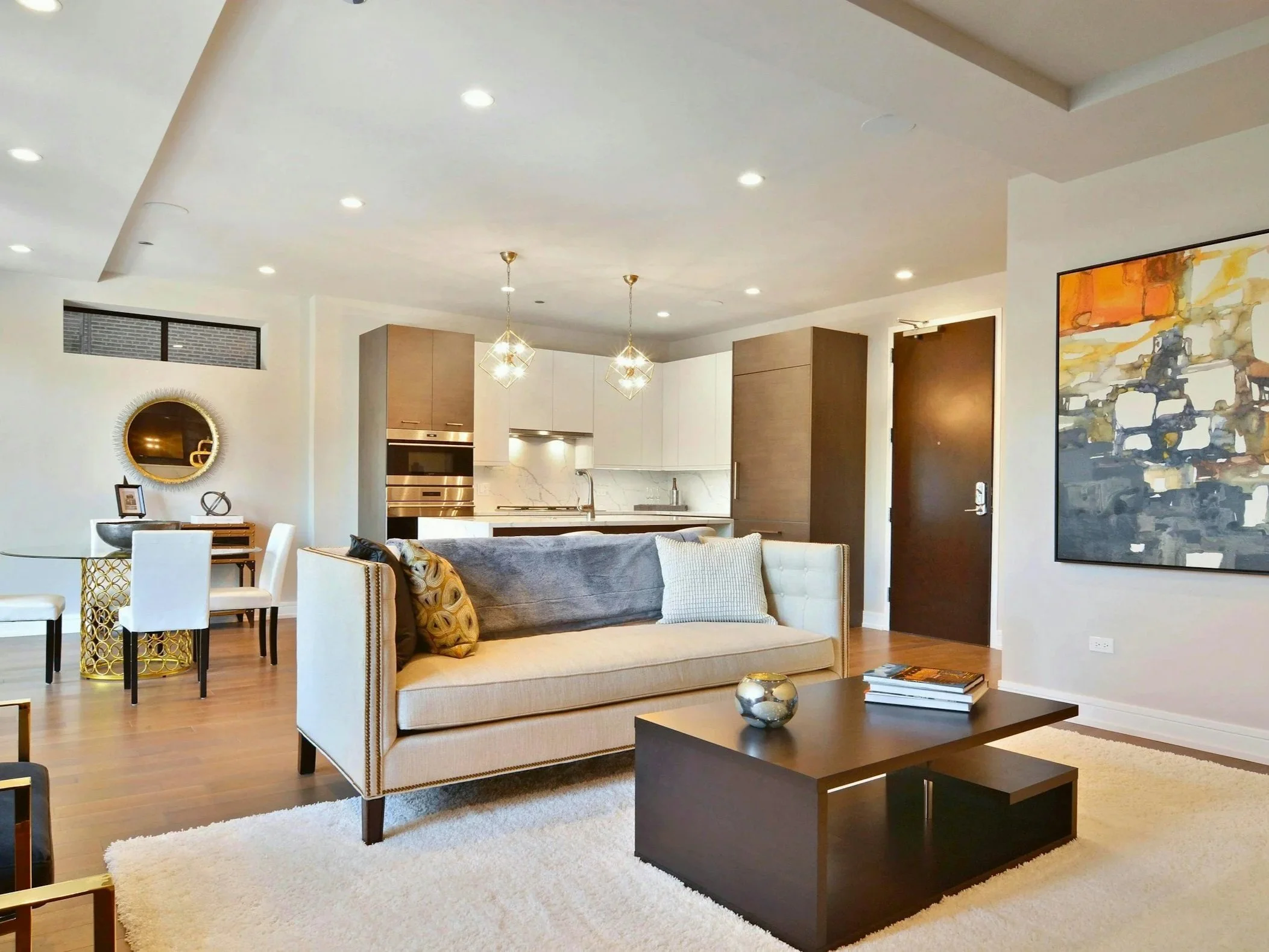 Modern living room with white sofa, dark wood coffee table, and abstract wall art, open to a kitchen with white cabinets and wooden accents