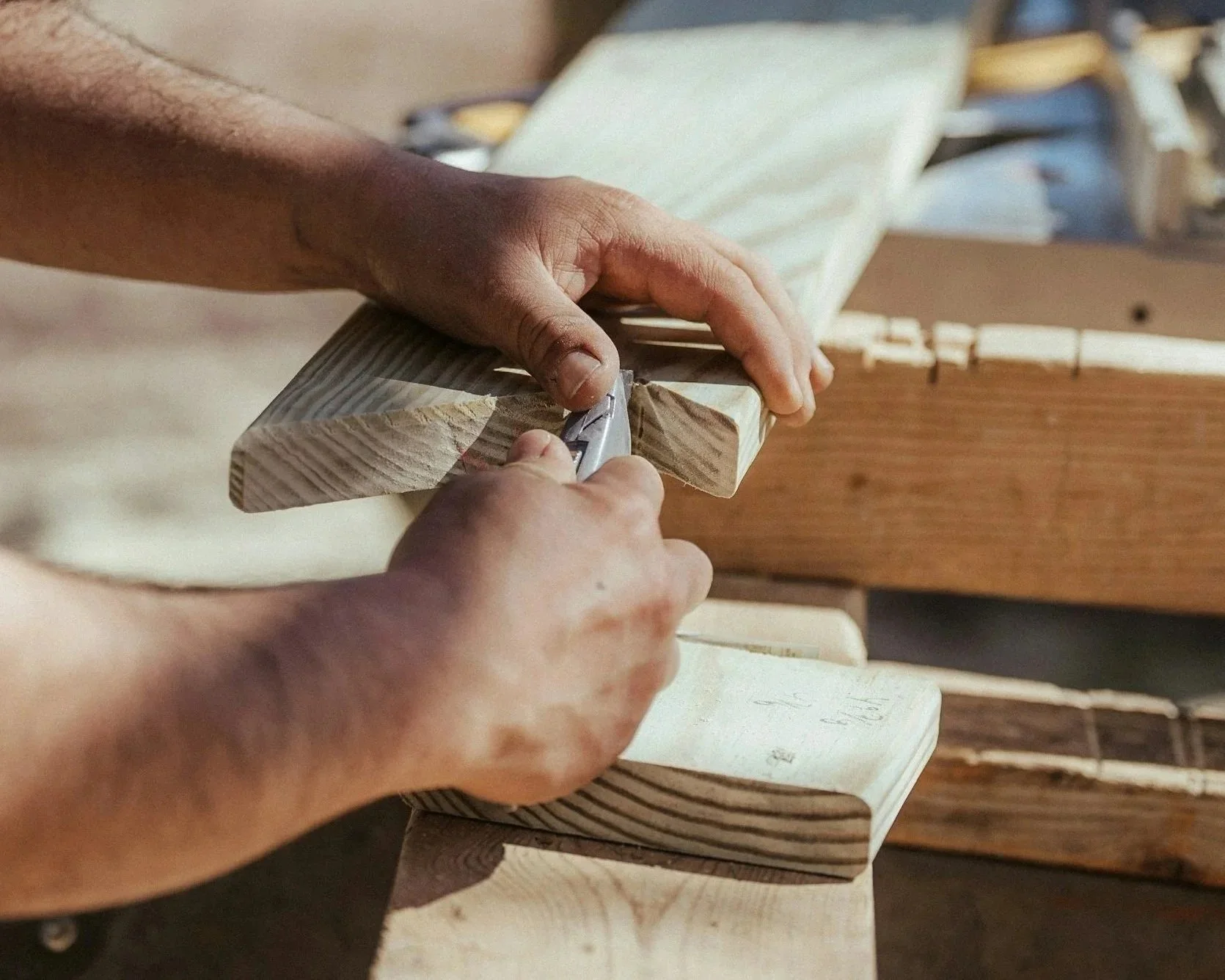 Close-up of a person's hands using a utility knife to cut a piece of wood, with wooden planks and construction materials in the background.